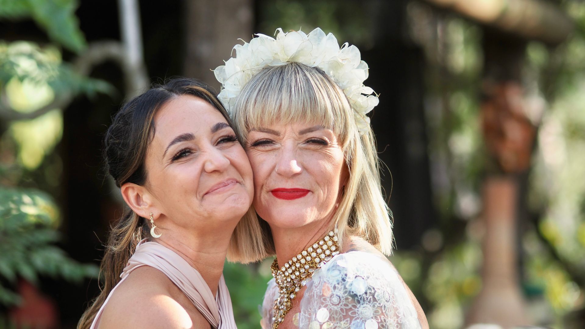 Two smiling women embrace outdoors, one wearing a white floral headpiece and dress.