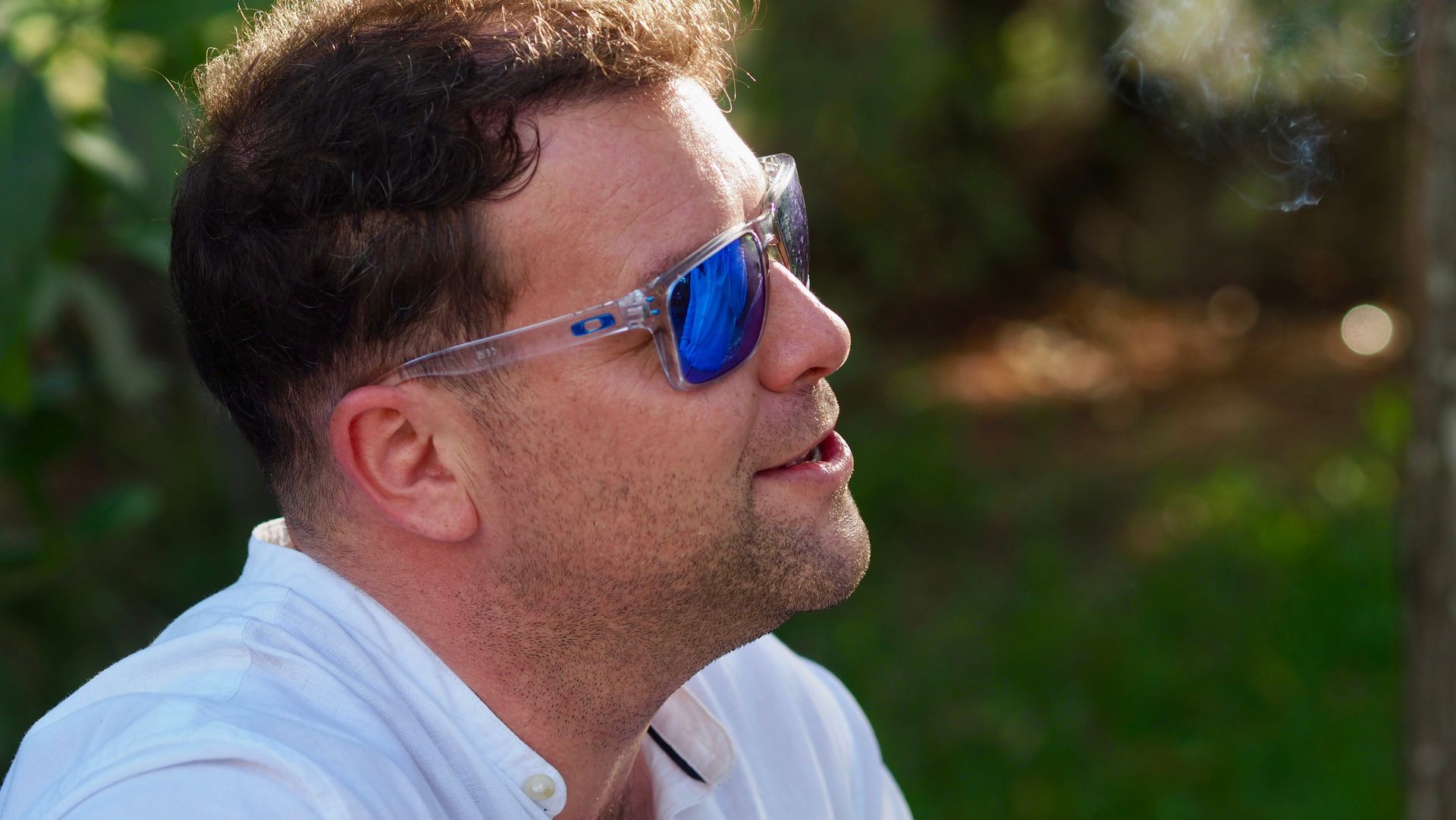 Man wearing blue mirrored sunglasses outdoors, looking upward in profile.