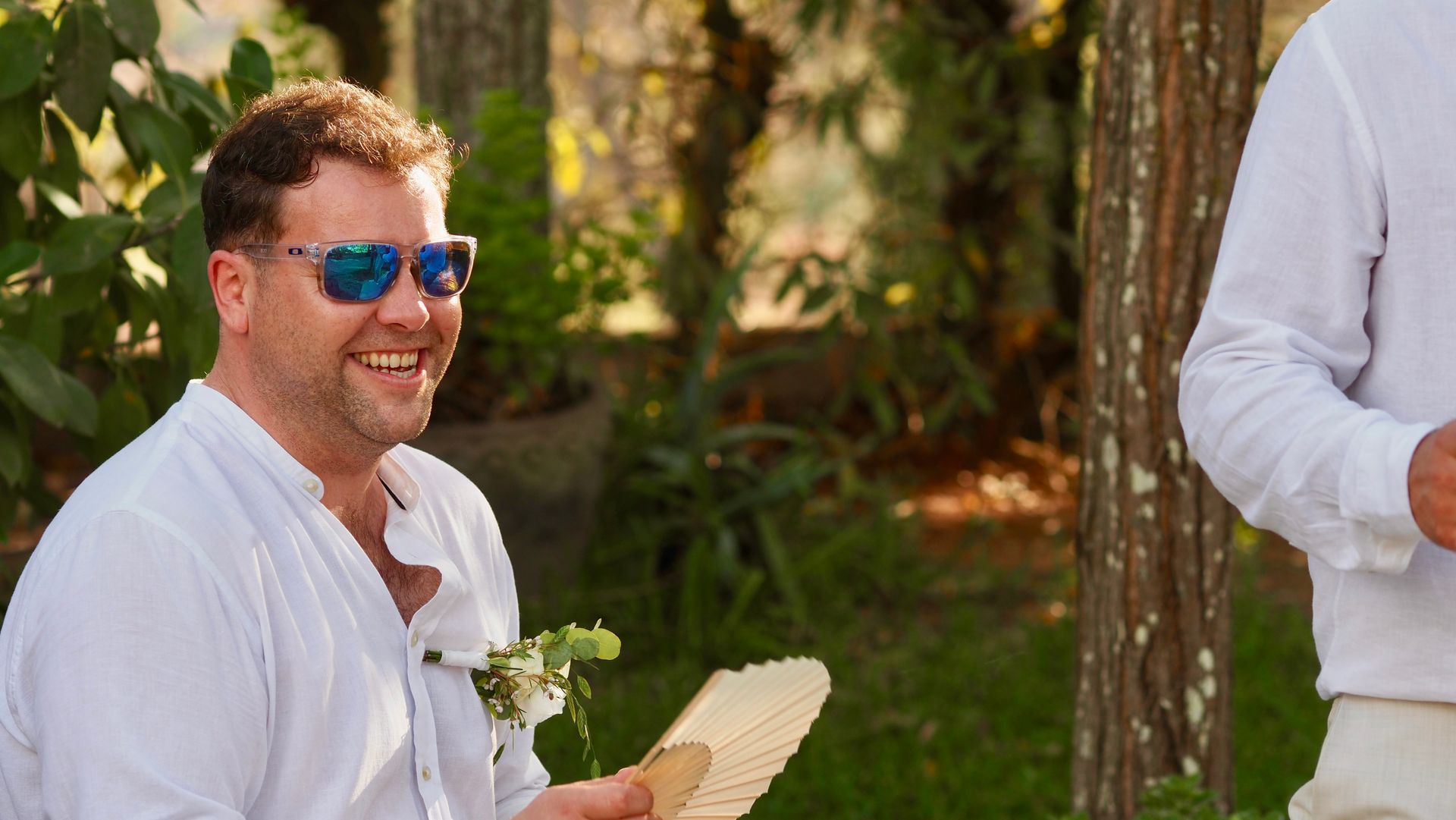 Smiling man in blue sunglasses holding a folded fan outdoors in a sunny garden setting