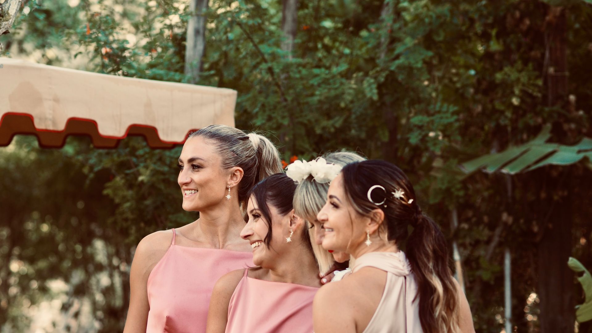 Three women in pink dresses posing outdoors in front of trees, smiling in profile.