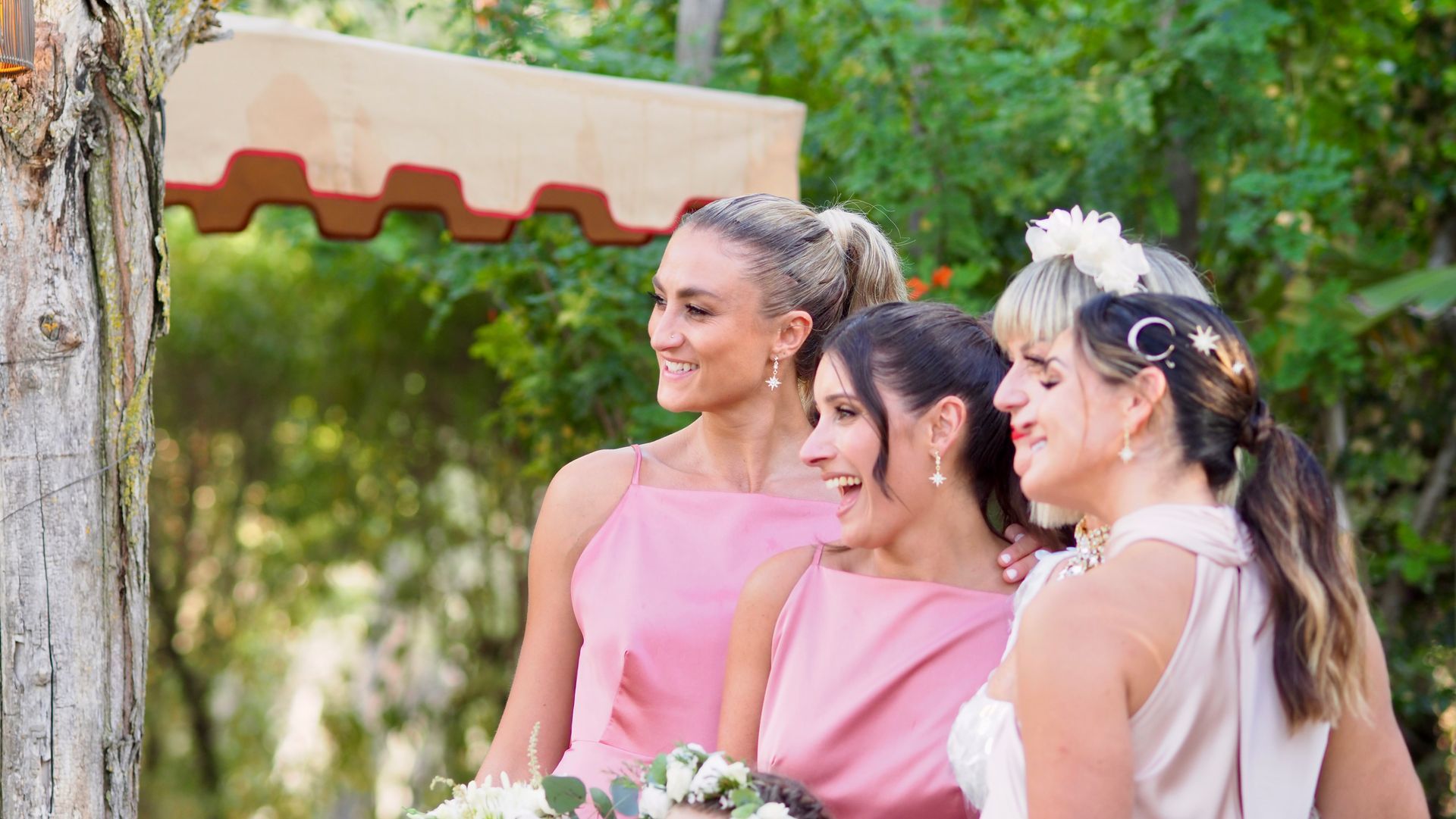 Three women in pink dresses pose together outdoors at a celebration, holding bouquets and smiling.