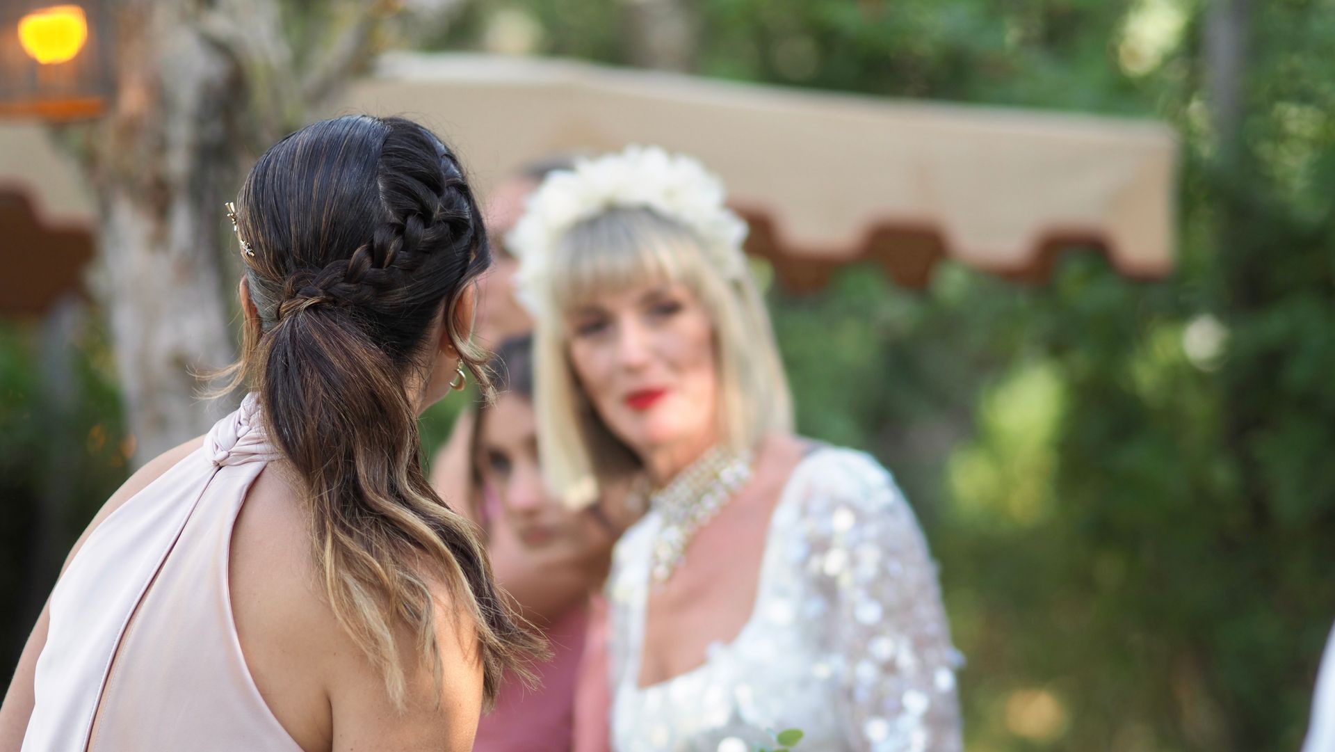 Two women in a garden, one facing away while the other in a white dress looks on with a blurred background.