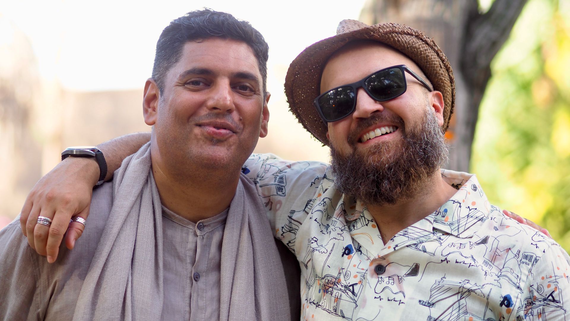 Two smiling friends with arms around each other outdoors, one wearing sunglasses and a hat