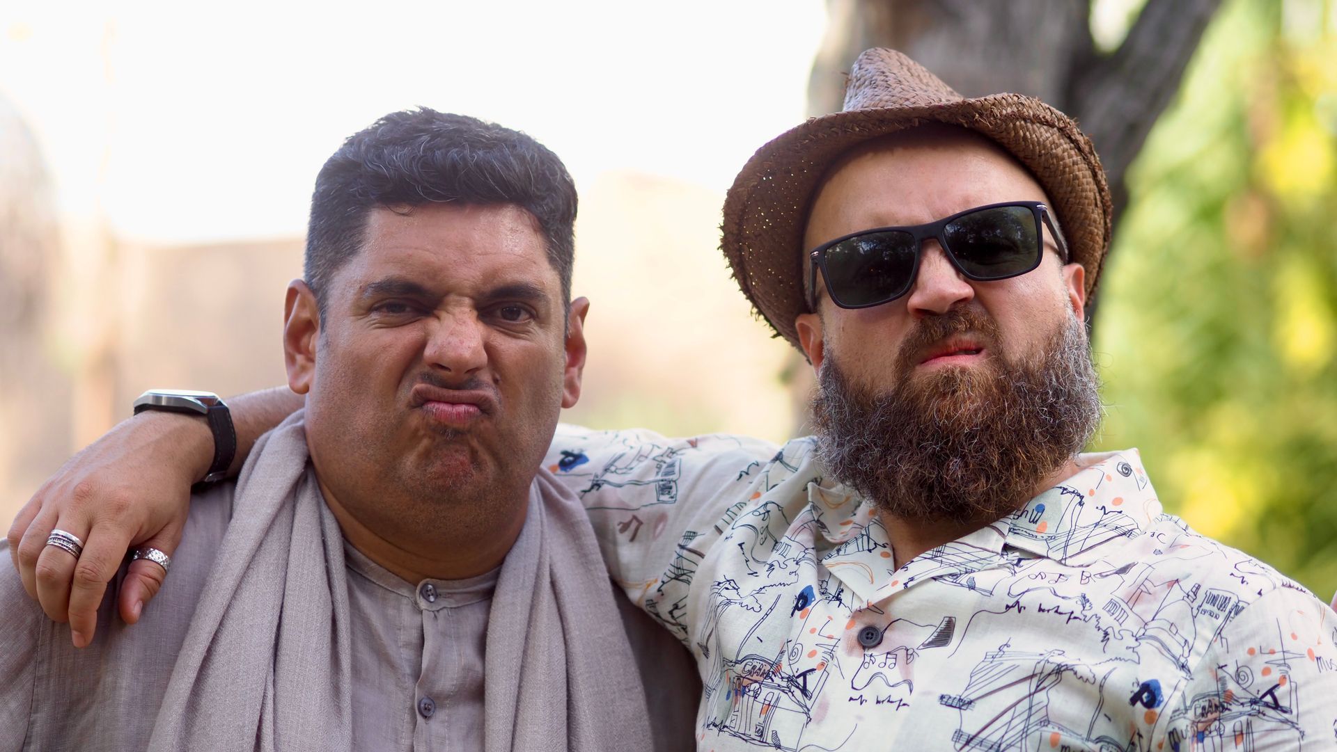 Two men pose arm-in-arm outdoors, one wearing sunglasses and a hat.