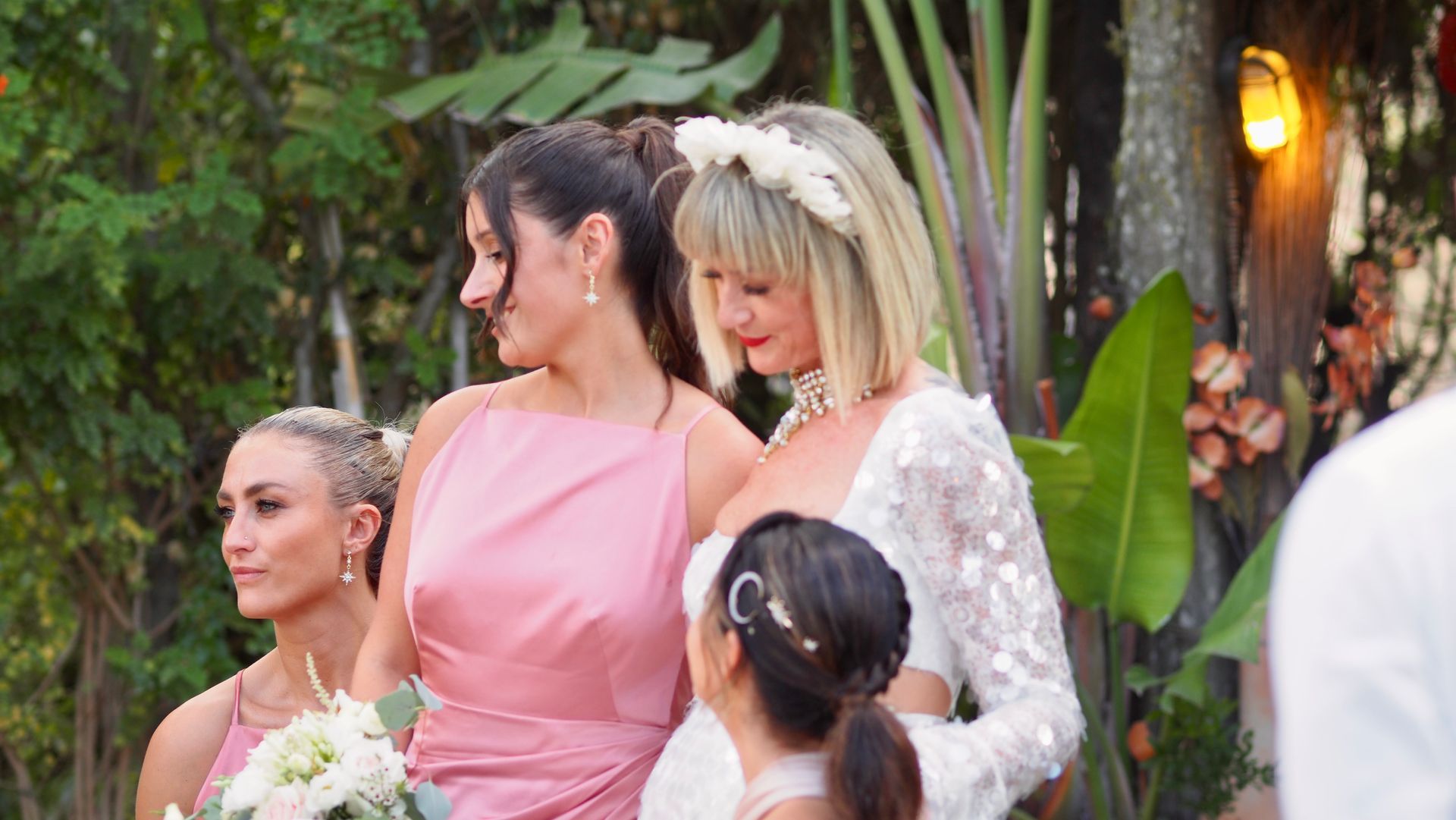 Four women in pastel dresses pose outdoors at a wedding, surrounded by tropical greenery.