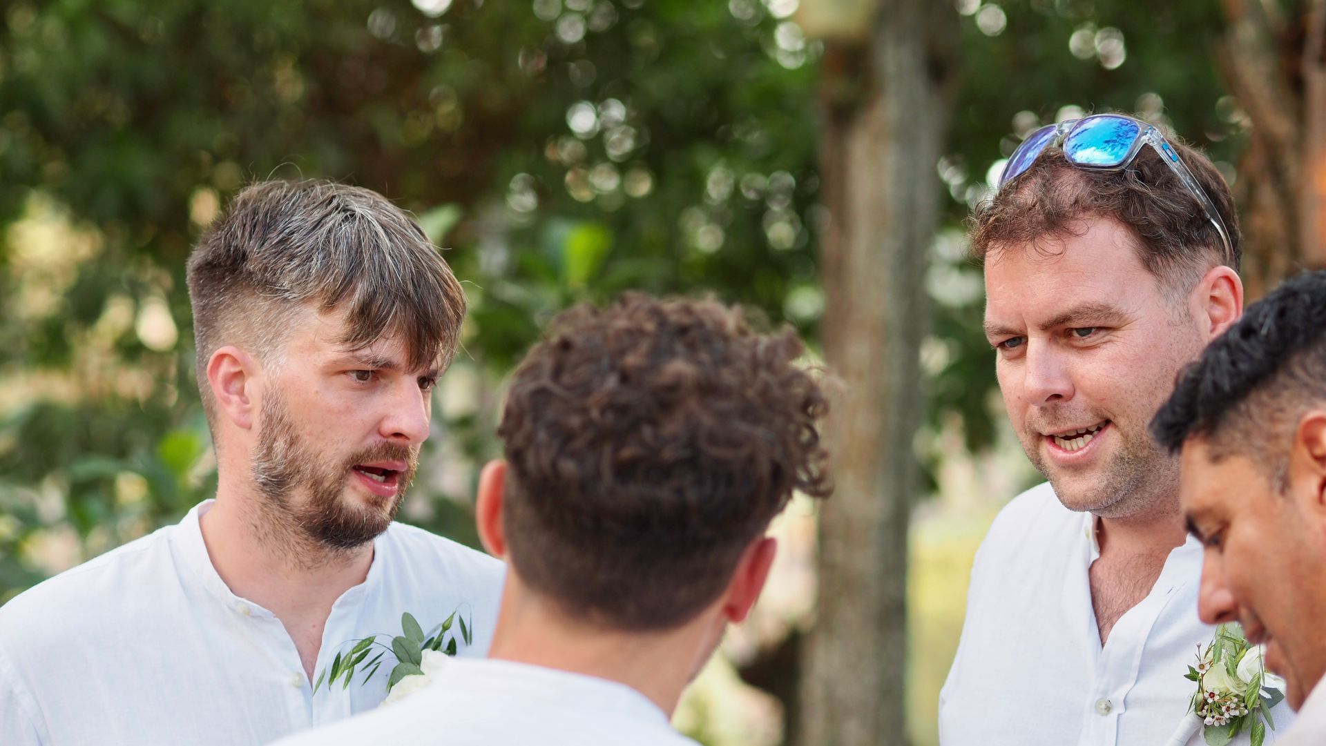 Three men in white shirts talking outdoors at a social gathering, with greenery in the background