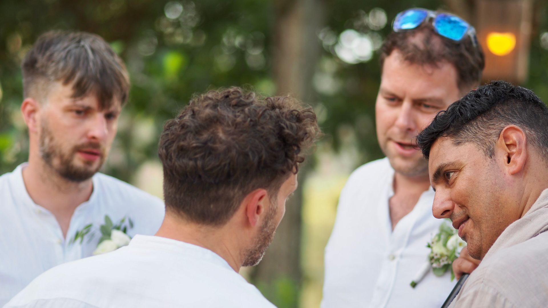 Four men in white shirts talking outdoors at a social event, with greenery in the background
