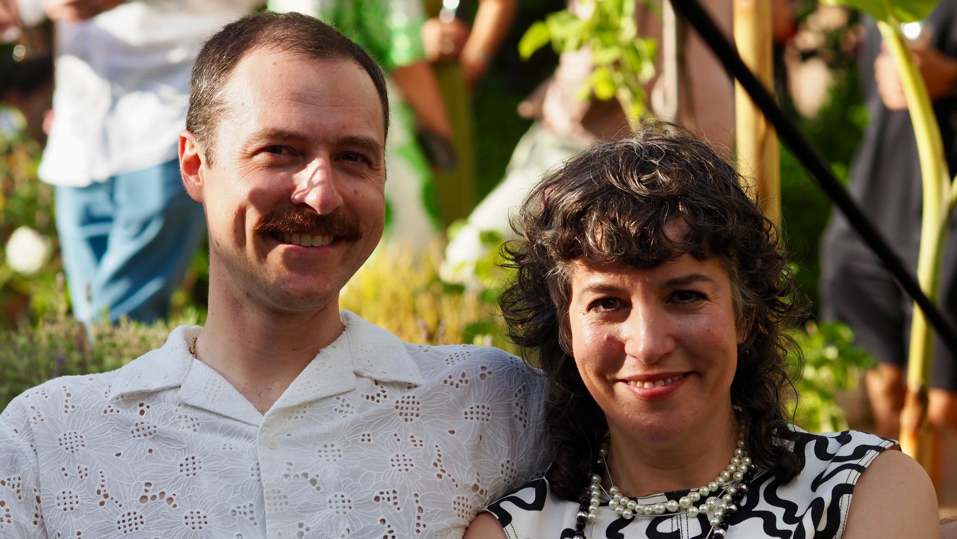 Two people smiling outdoors in a garden, with flowers and sunlight in the background.