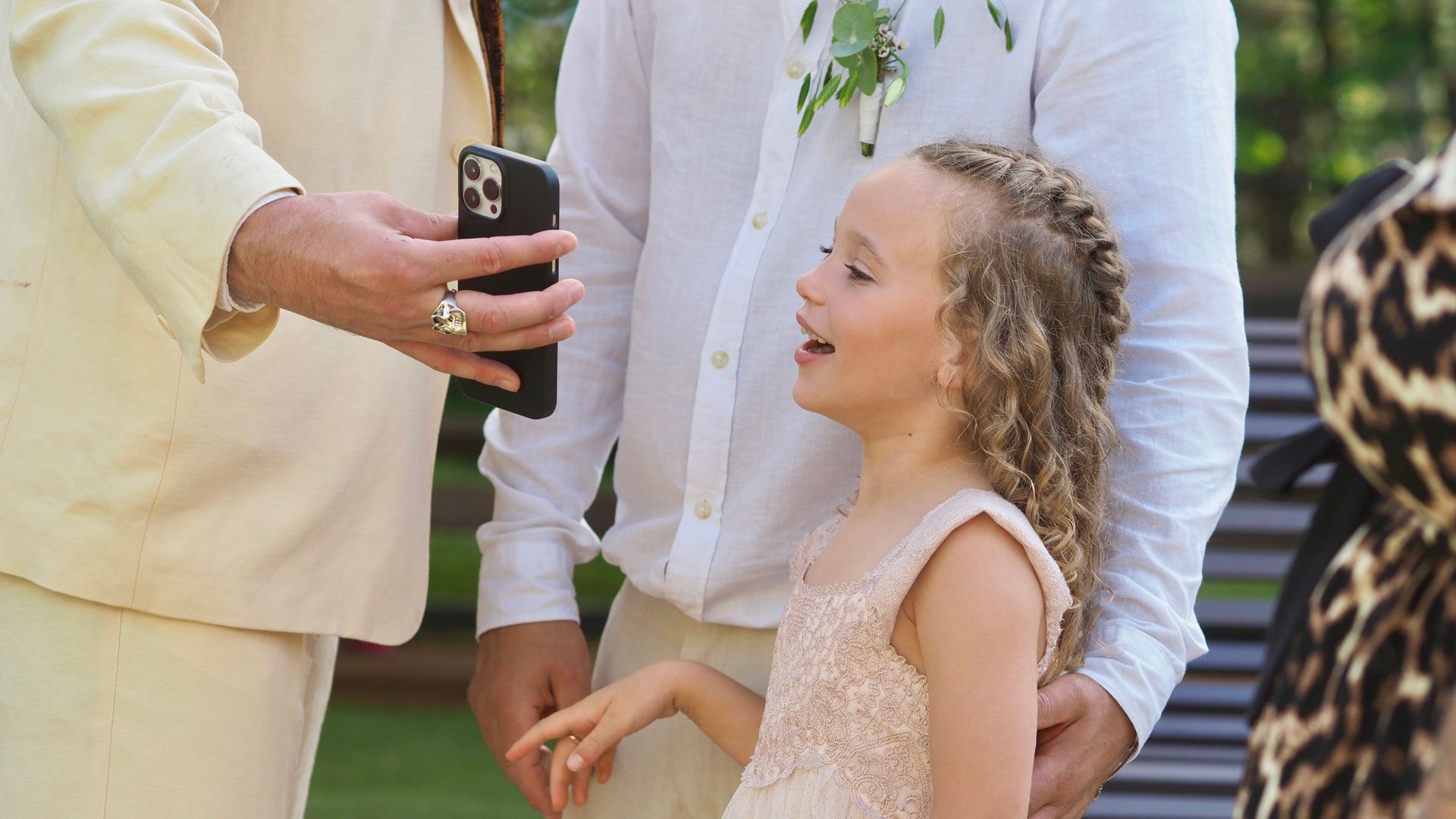 Child smiling at a formal outdoor event while an adult shows a phone nearby