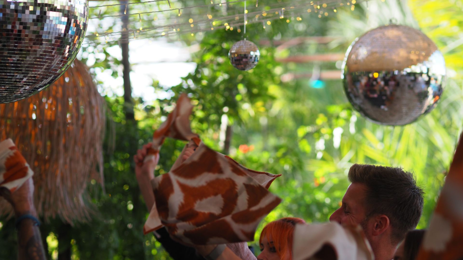 People dancing outdoors under hanging silver disco balls in a lush, sunlit garden setting