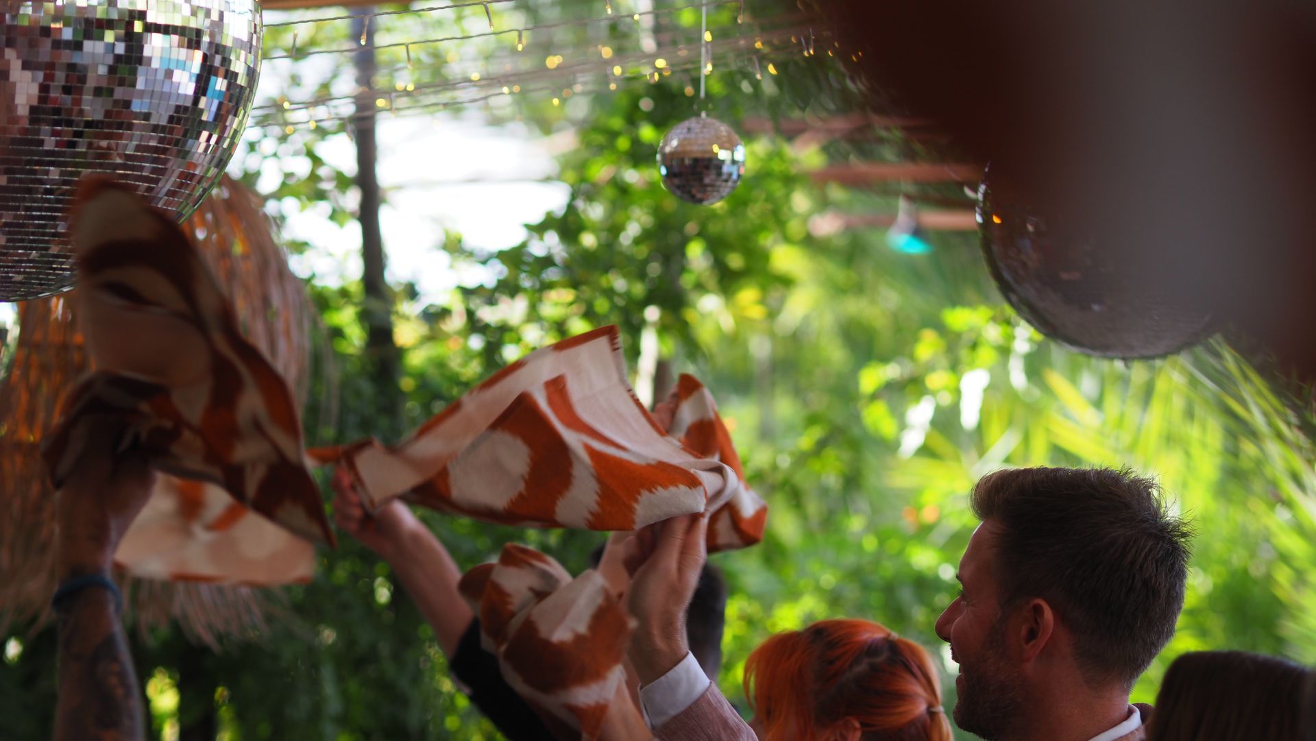 People waving orange-and-white cloths in a festive outdoor crowd, with greenery and hanging decorations.