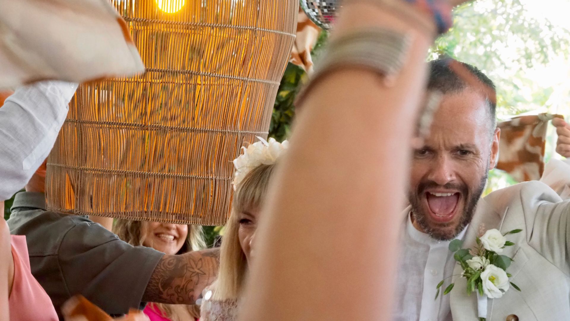 Smiling man in a white shirt raises his arms amid a lively celebration under a woven canopy