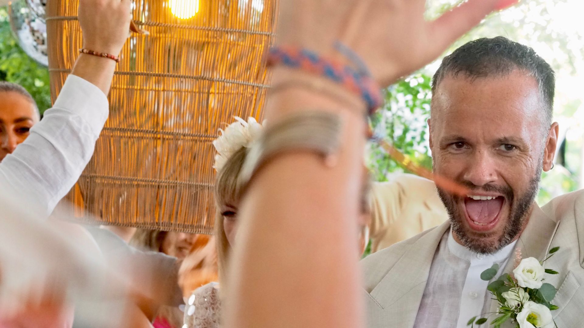 Smiling man in a white suit at a festive celebration, surrounded by blurred guests and warm lights