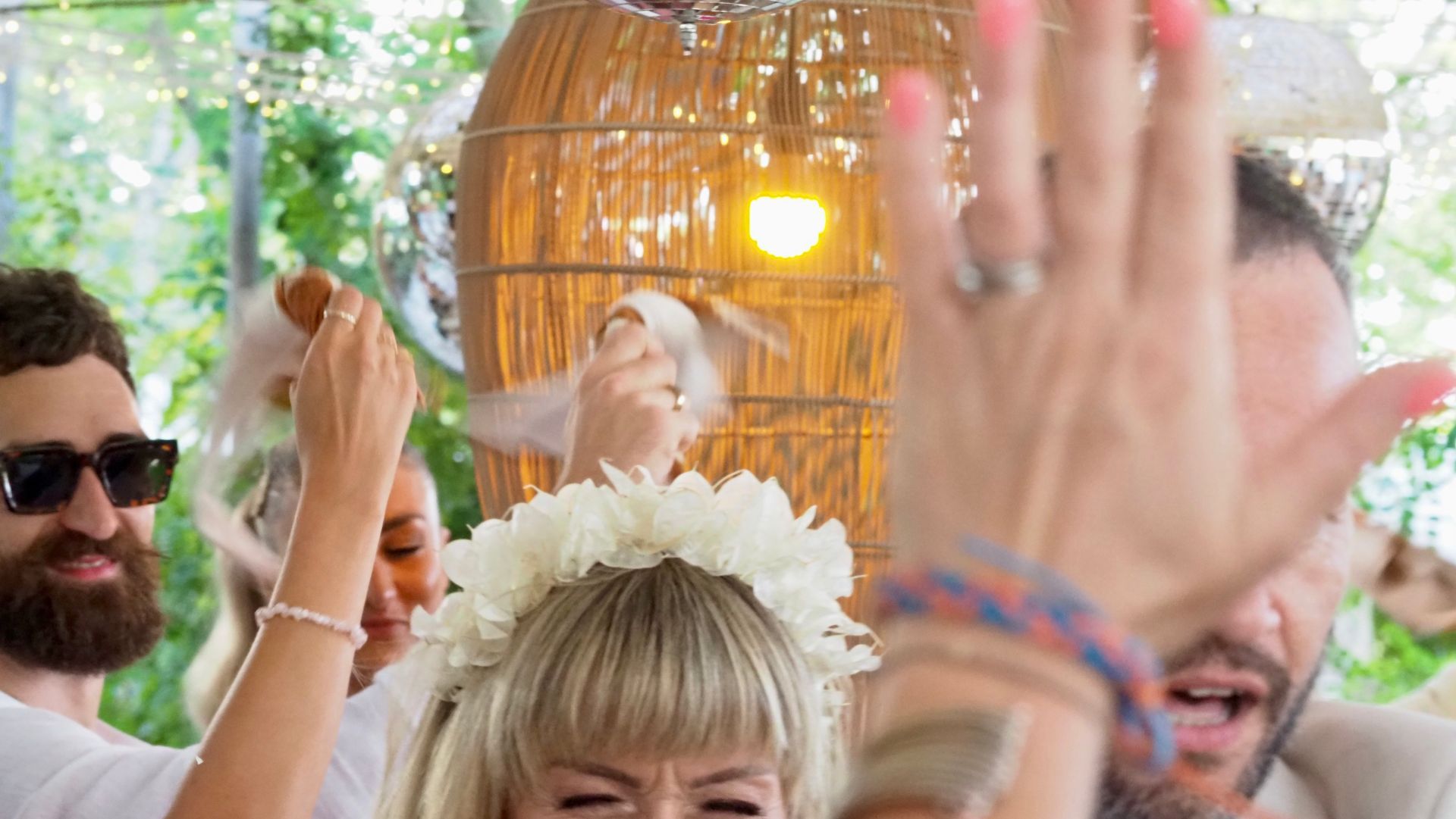 People raising hands at a lively outdoor party, with a woven lantern and sunlight in the background