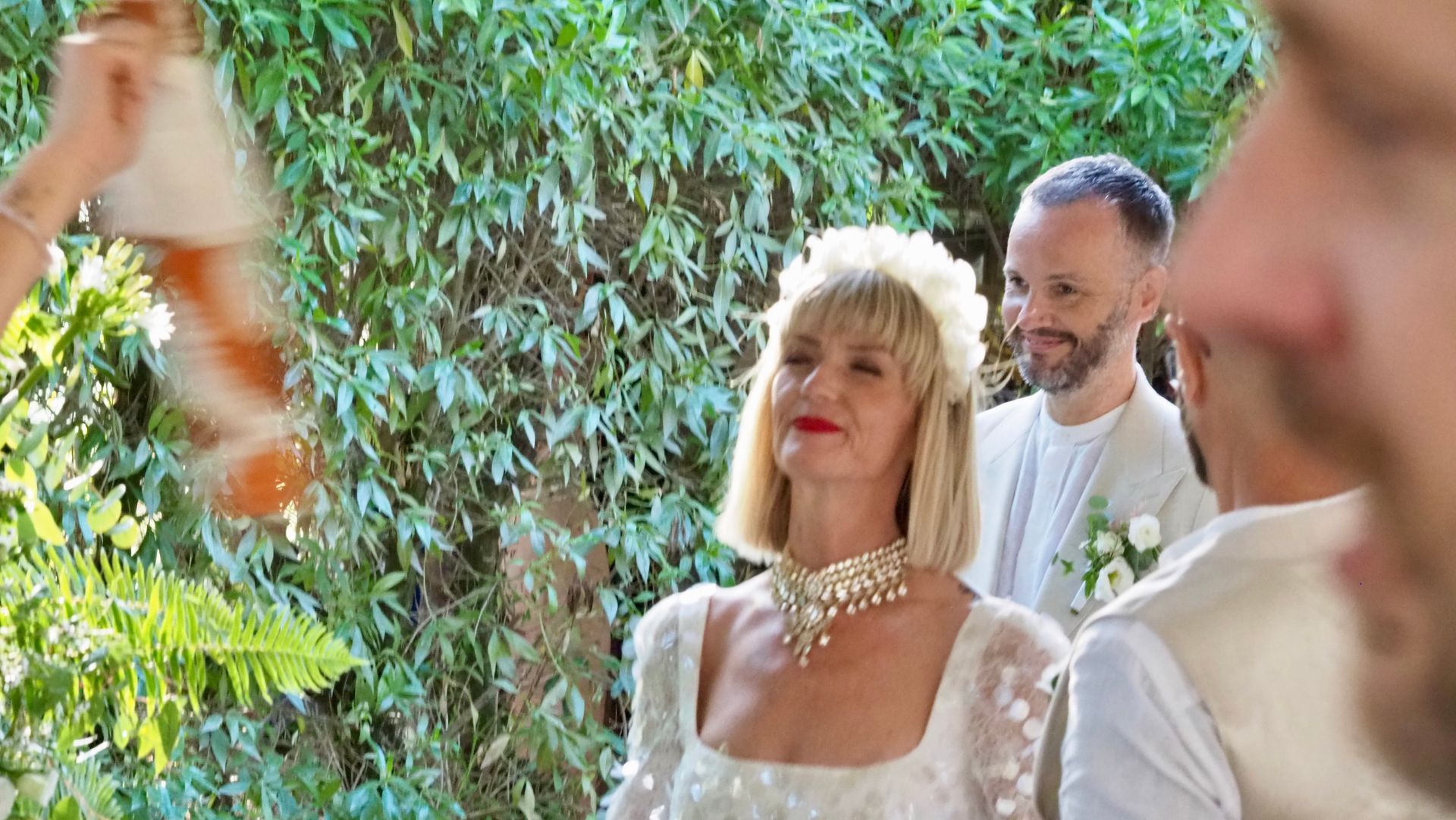 Smiling woman in white dress and pearl necklace at an outdoor gathering with greenery behind her
