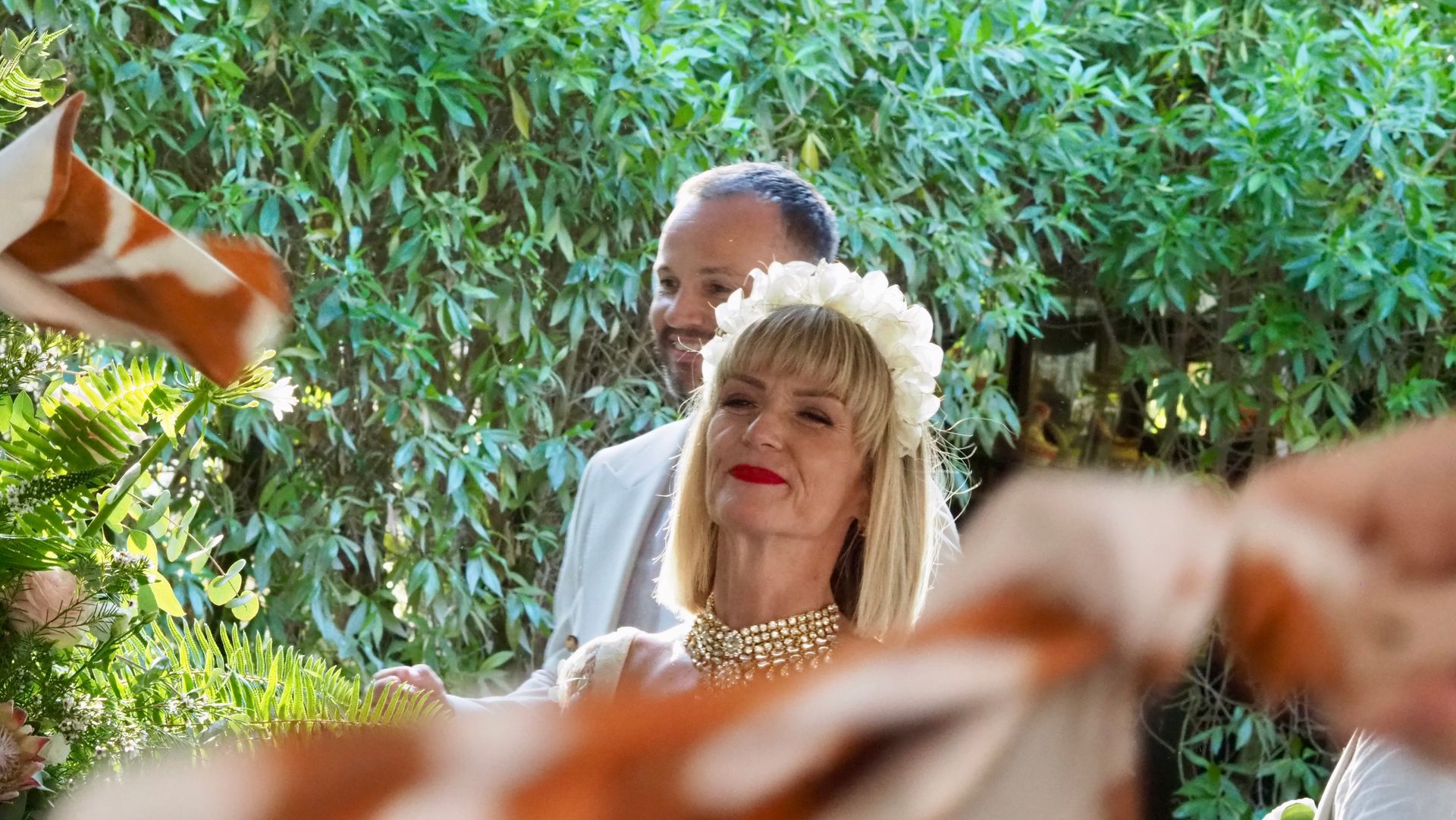 Blonde bride in white dress and veil standing outdoors amid greenery and blurred ribbons