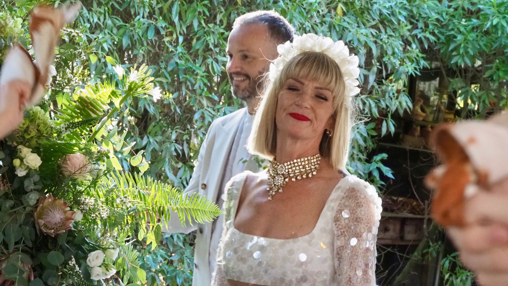 Two people posing outdoors in a garden, with lush greenery and flowers in the foreground
