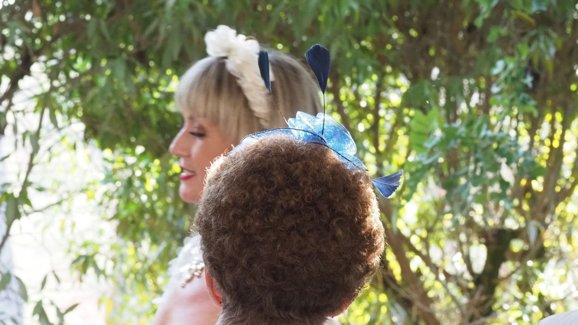 Two people in a leafy outdoor setting, one with a blue floral headpiece and one with a furry brown hat