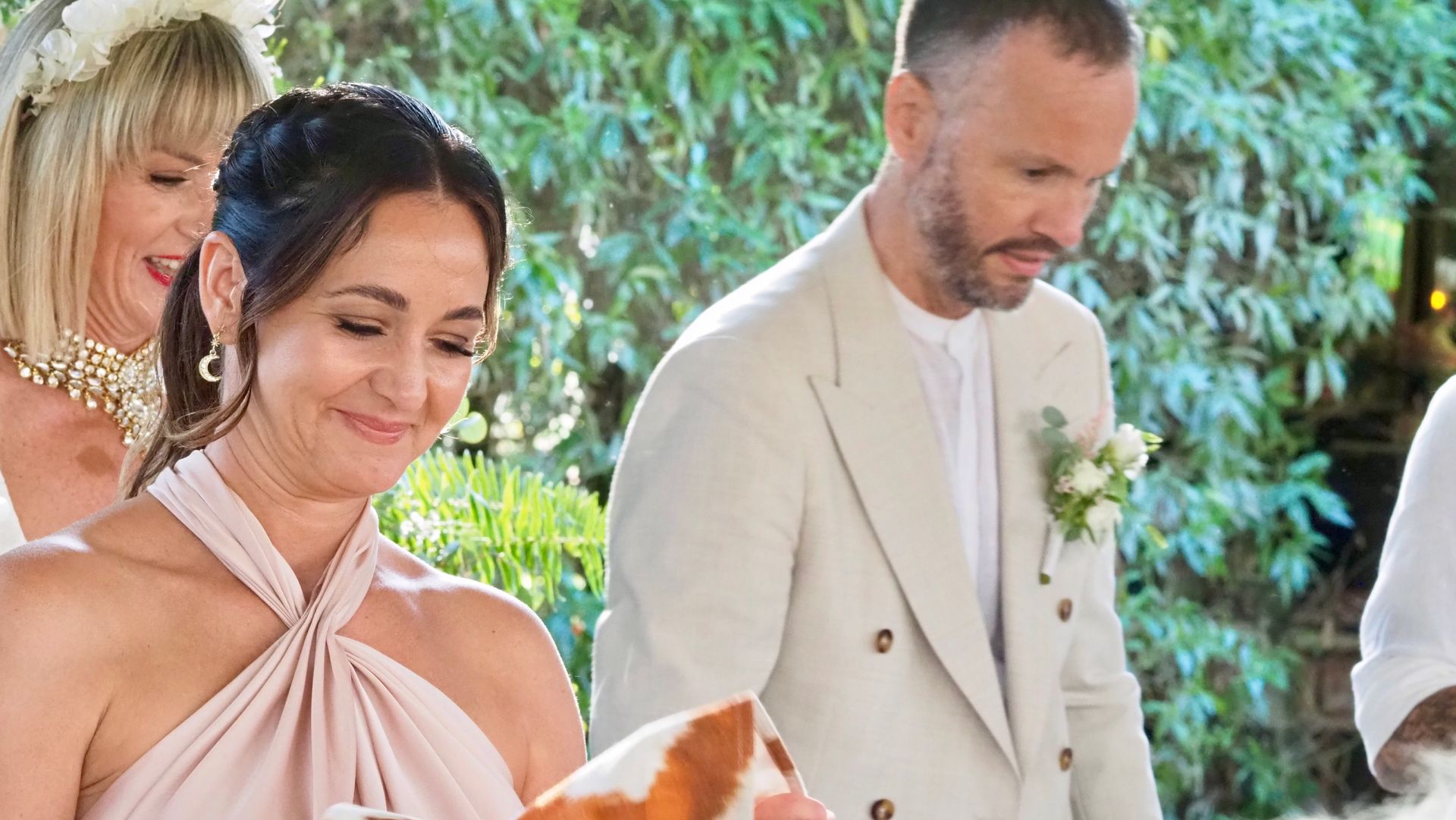 People in formal attire standing outdoors at a wedding ceremony, smiling near a floral arch.