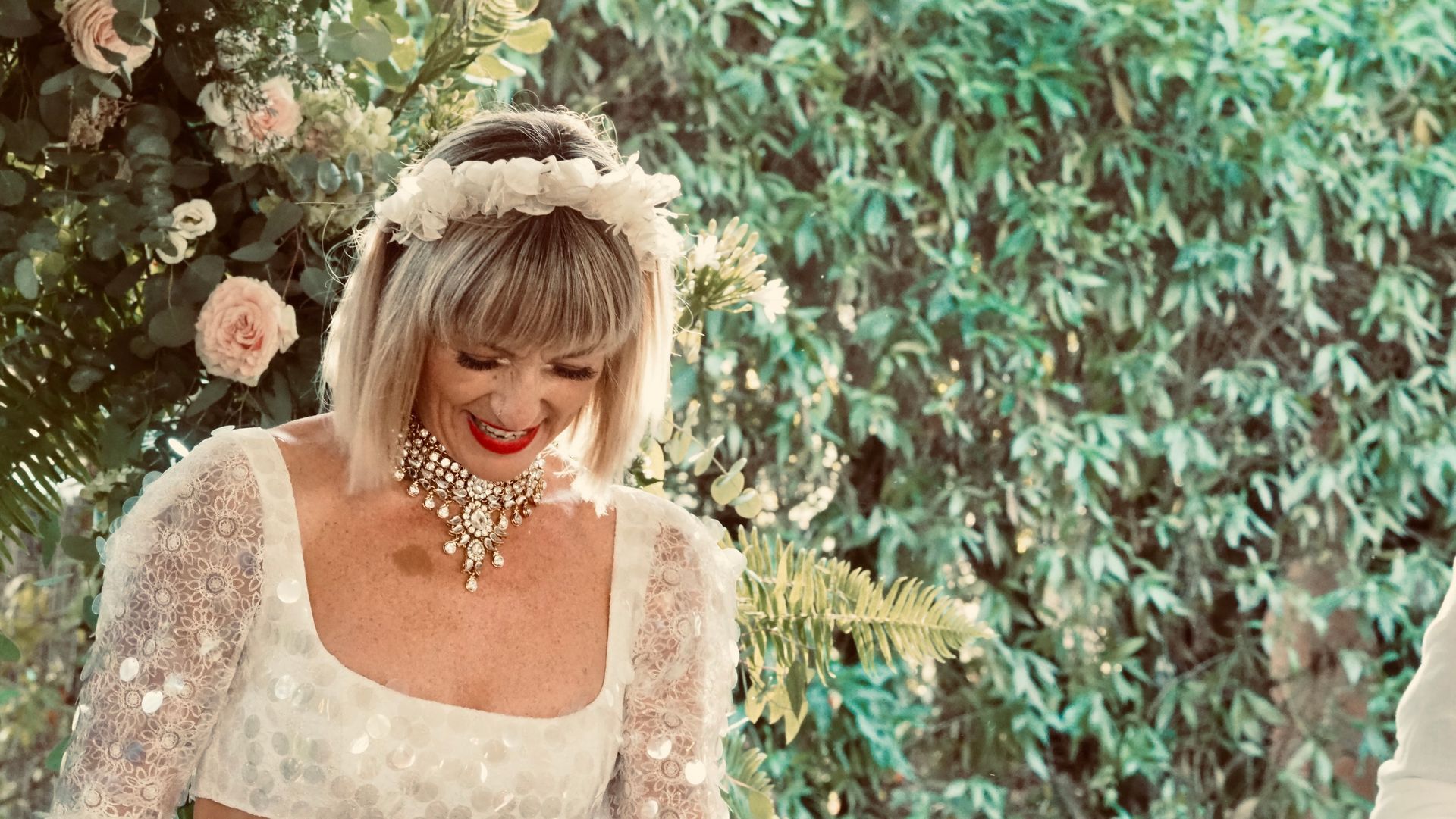 Woman in a white dress and jeweled necklace smiling beside a flower-covered backdrop