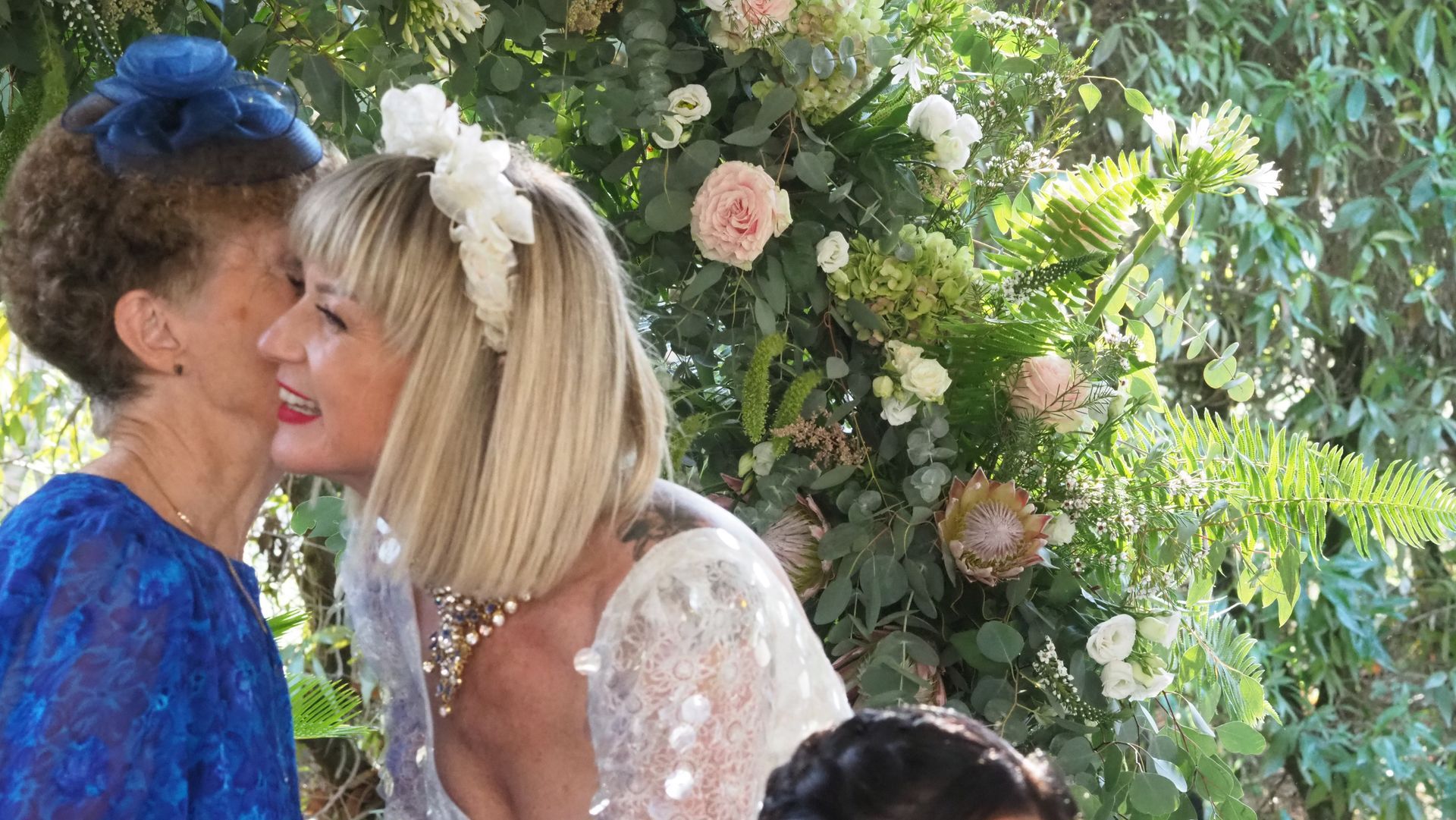 Two women greet with a cheek kiss in a garden, one in blue and one in white amid flowers.