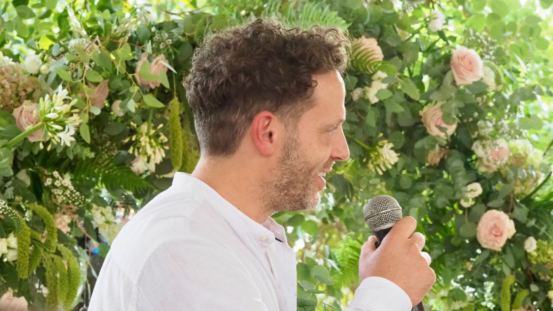 Man speaking into a microphone in front of a lush floral wall with pink and white roses