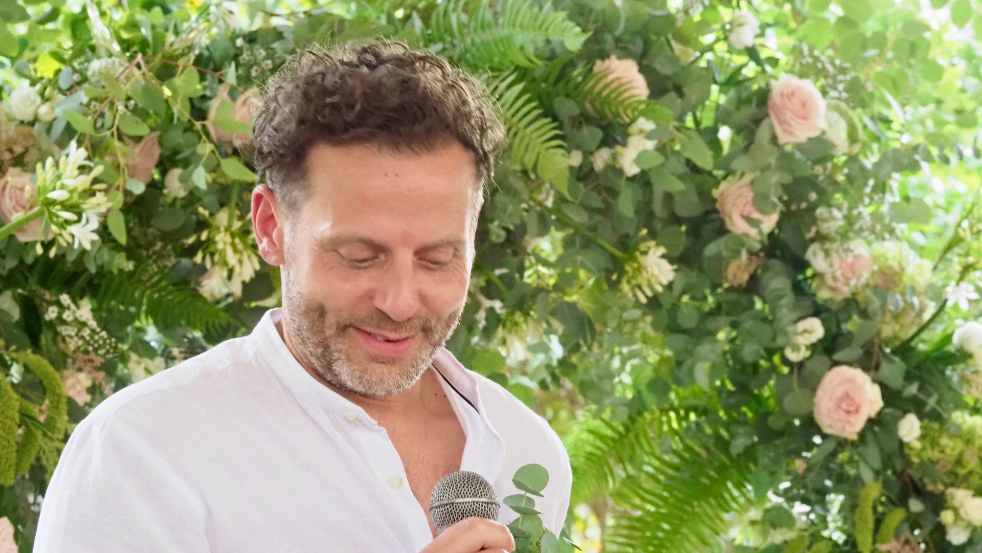 Man in white shirt holding a microphone in a lush flower-covered garden, eyes closed and smiling.