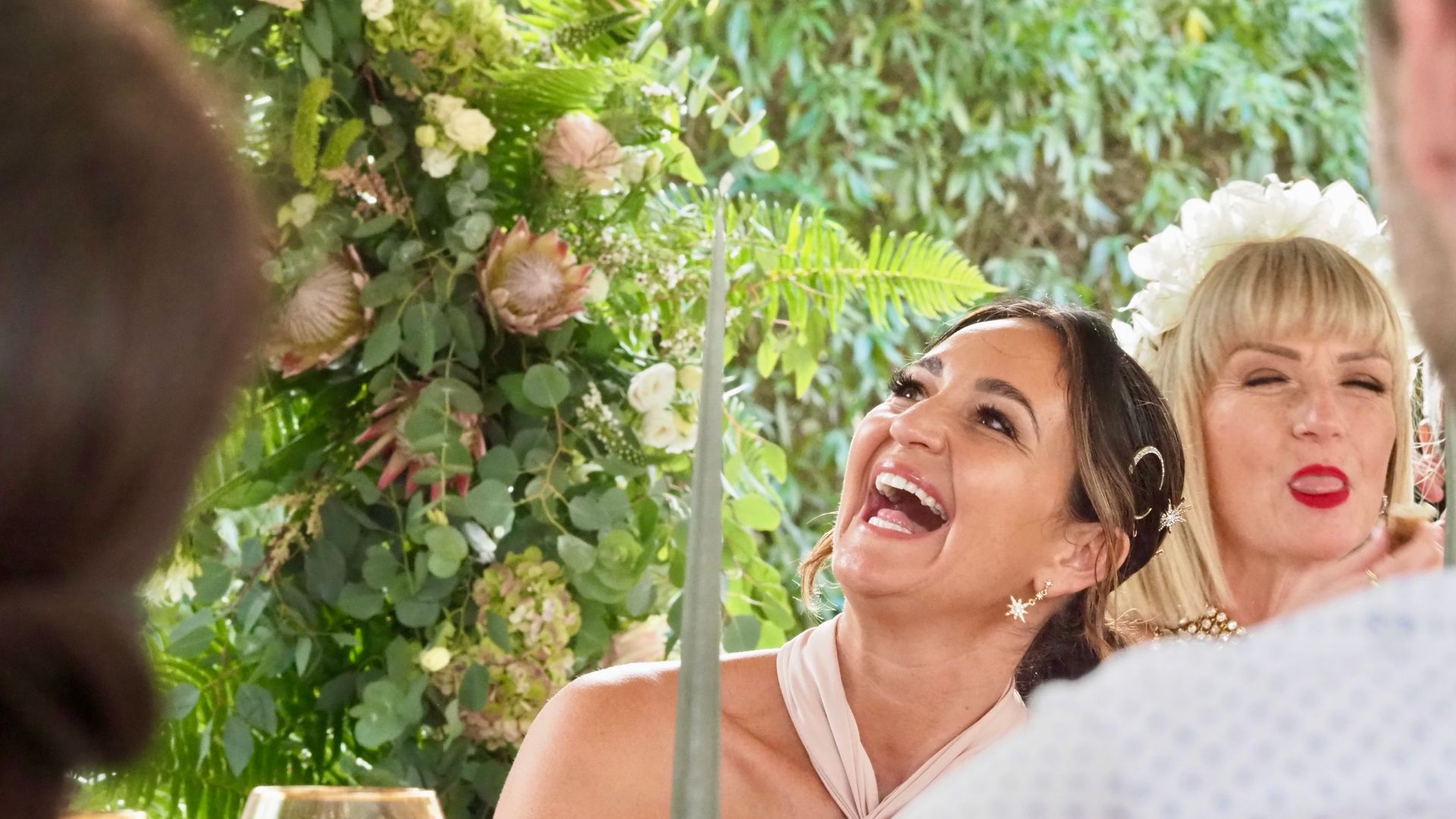 Two women smiling and looking up amid greenery and flowers at an outdoor celebration