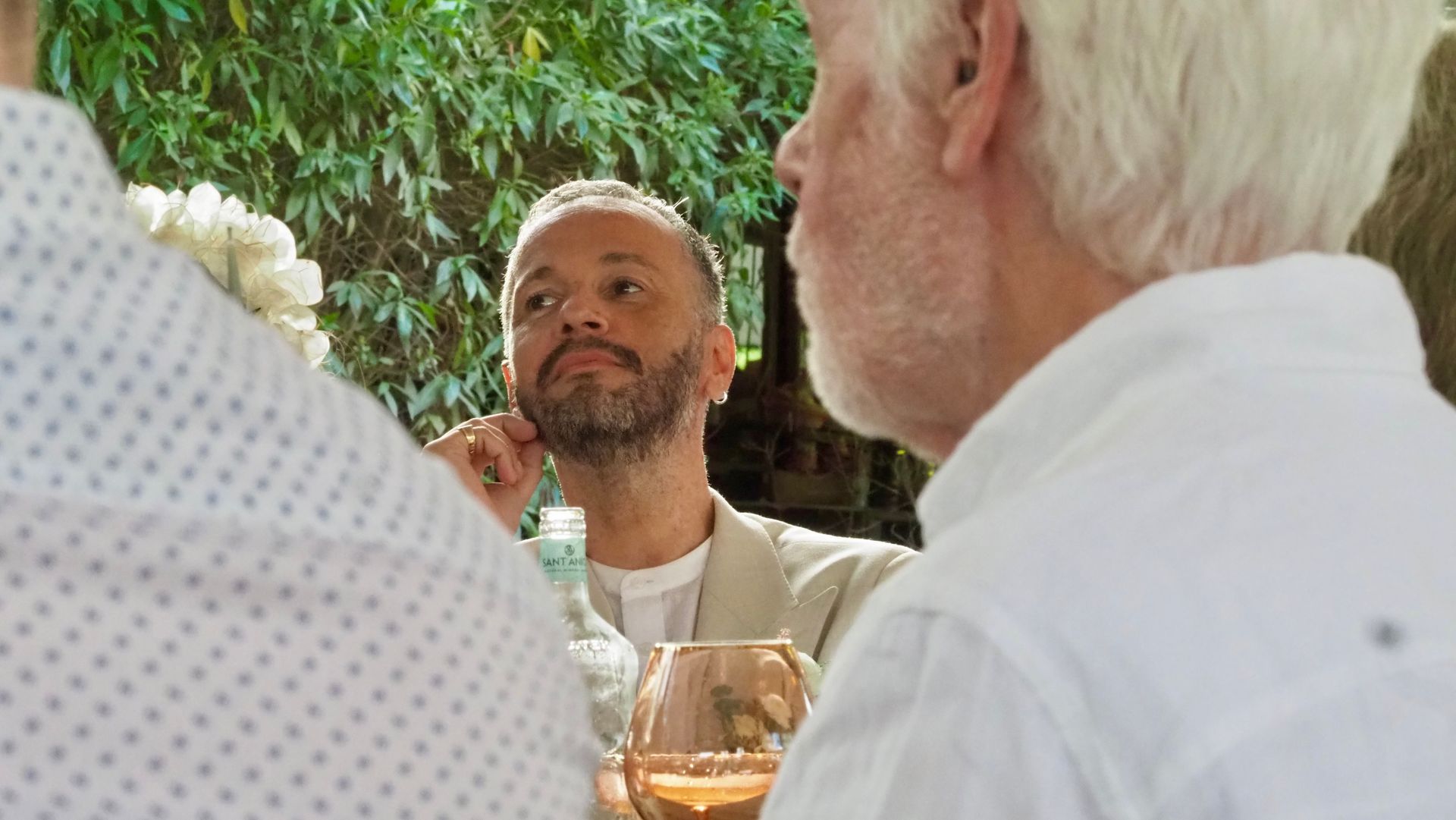 People seated outdoors in conversation, with a man holding a drink in the center.