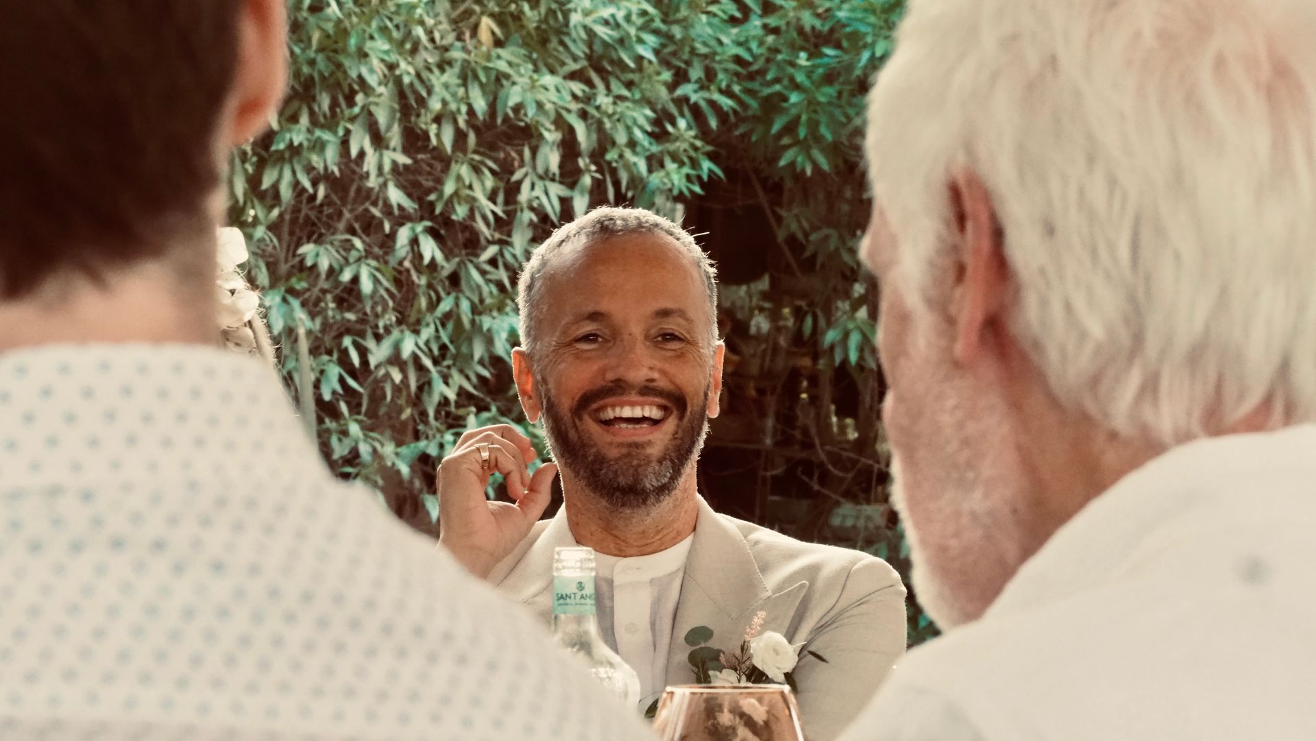 Smiling man in a light suit speaking at a formal gathering, seen between two guests in the foreground