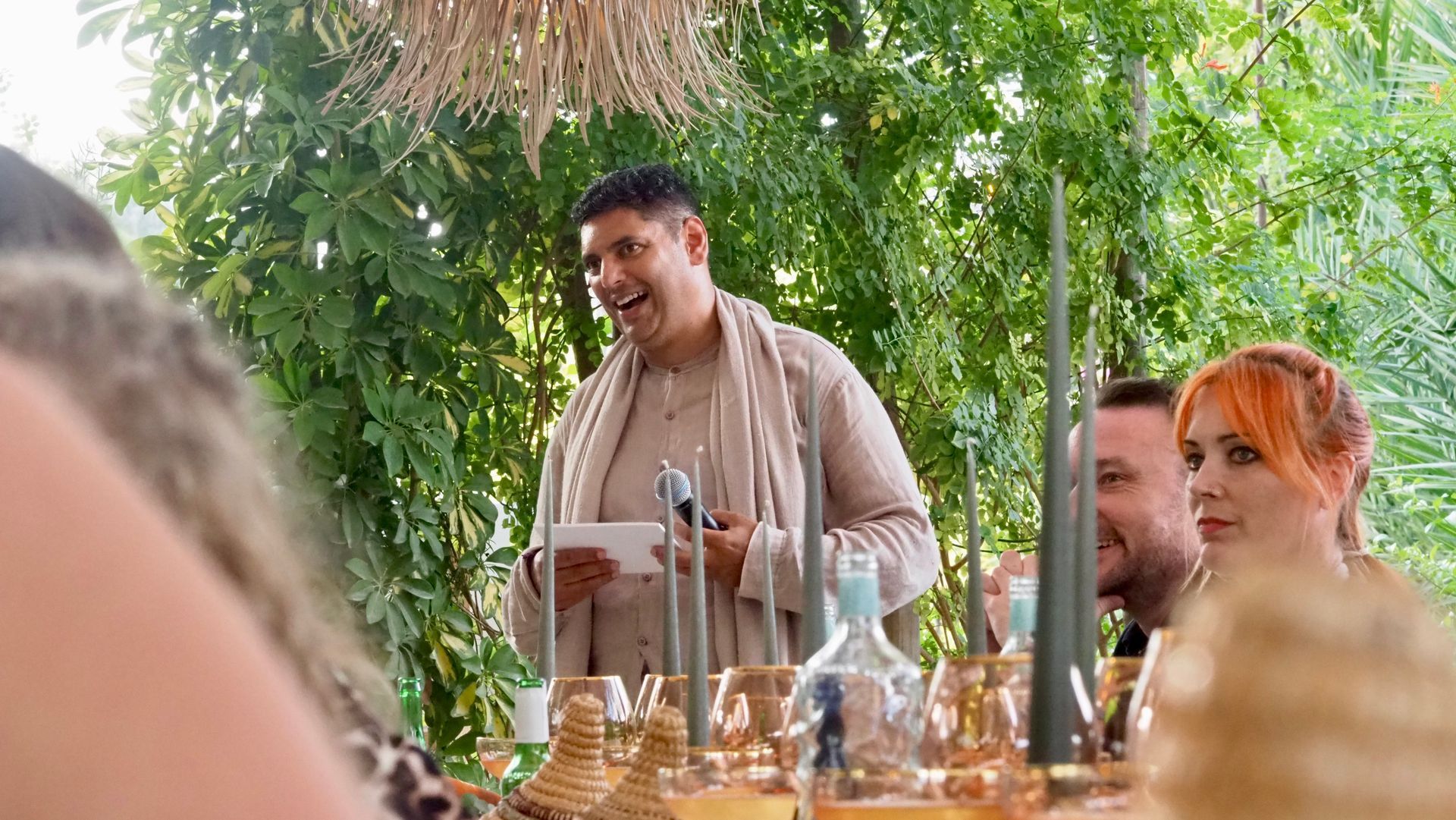 People seated at a decorated outdoor table, with a smiling speaker holding papers under green foliage.