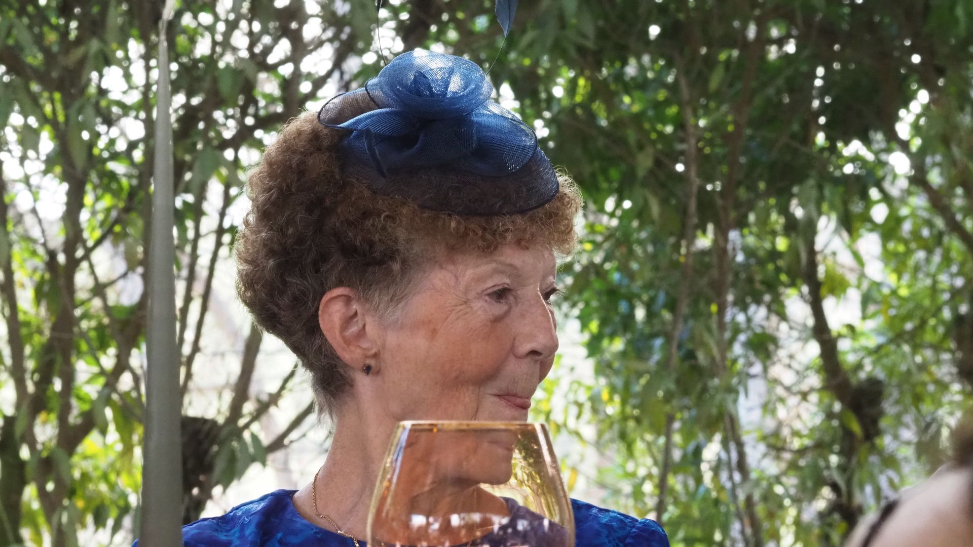 Person in a blue outfit and hat holding a drink, outdoors among trees.