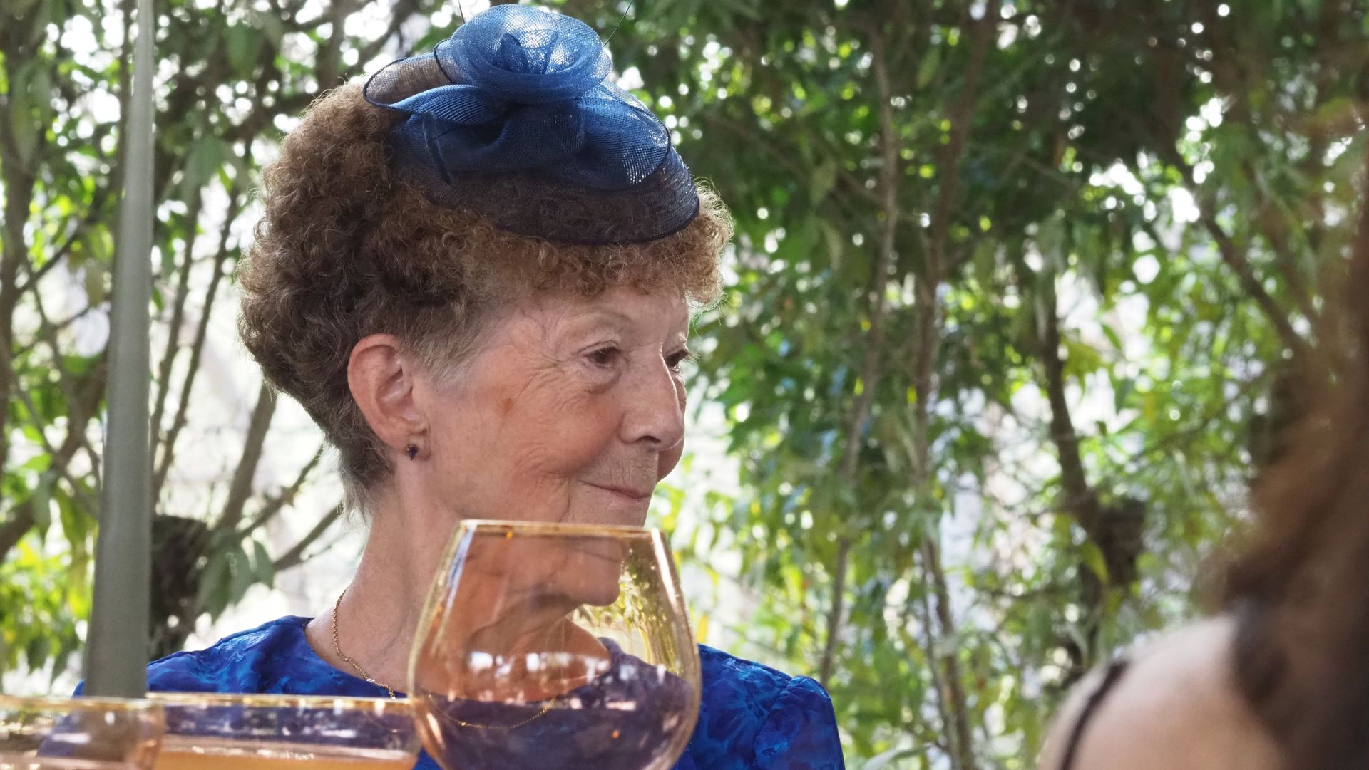 Woman in a blue dress and hat raises a glass at an outdoor gathering