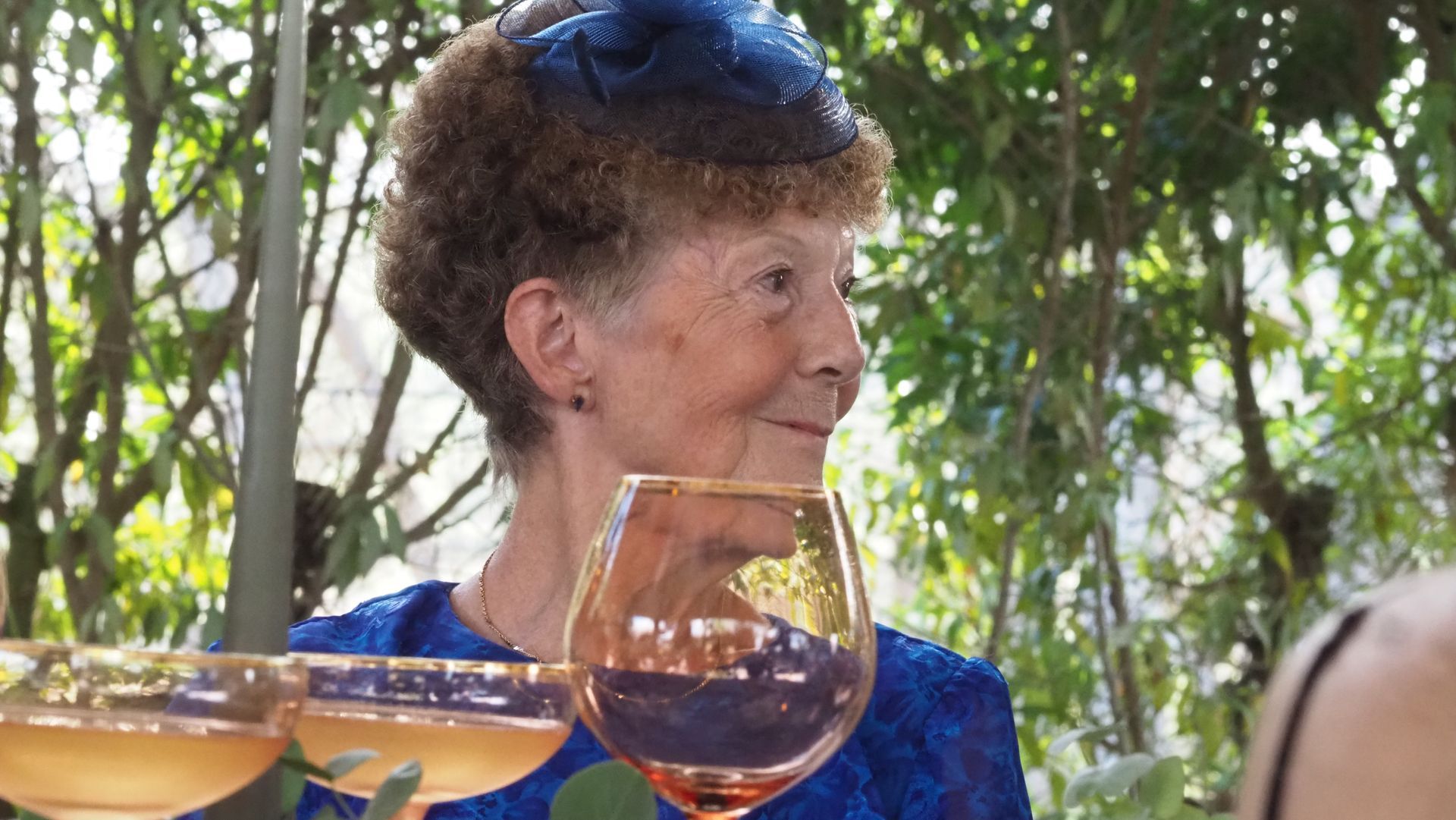 Woman in a blue outfit and hat holds a wine glass outdoors at a social gathering.