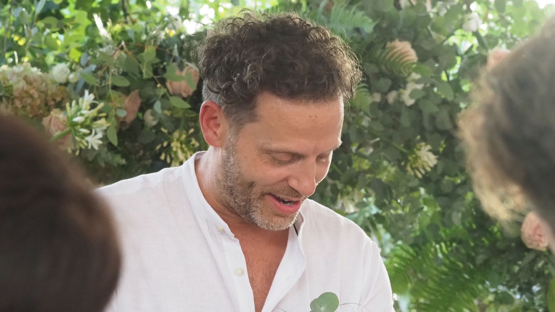 Smiling man in a white shirt outdoors, with leafy greenery in the background.
