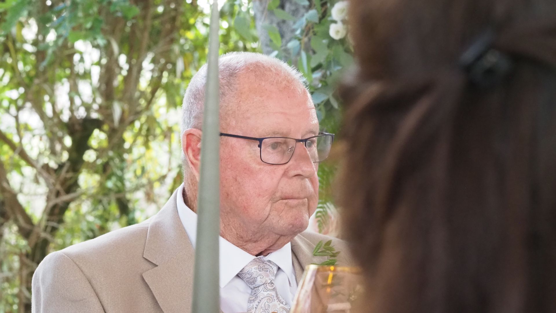Elderly man in a tan suit and glasses, seated outdoors, seen through a vertical pole in the foreground