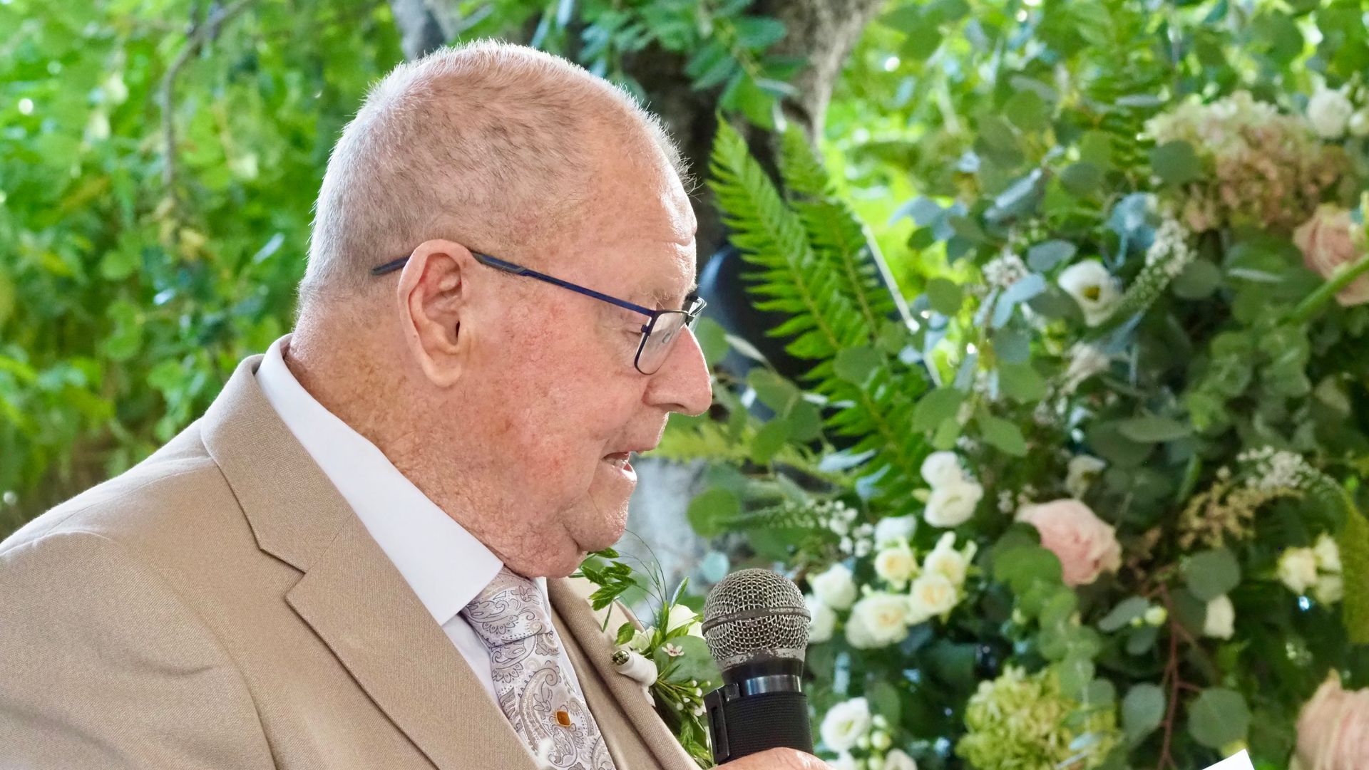 Man speaking into a microphone in front of a lush floral backdrop