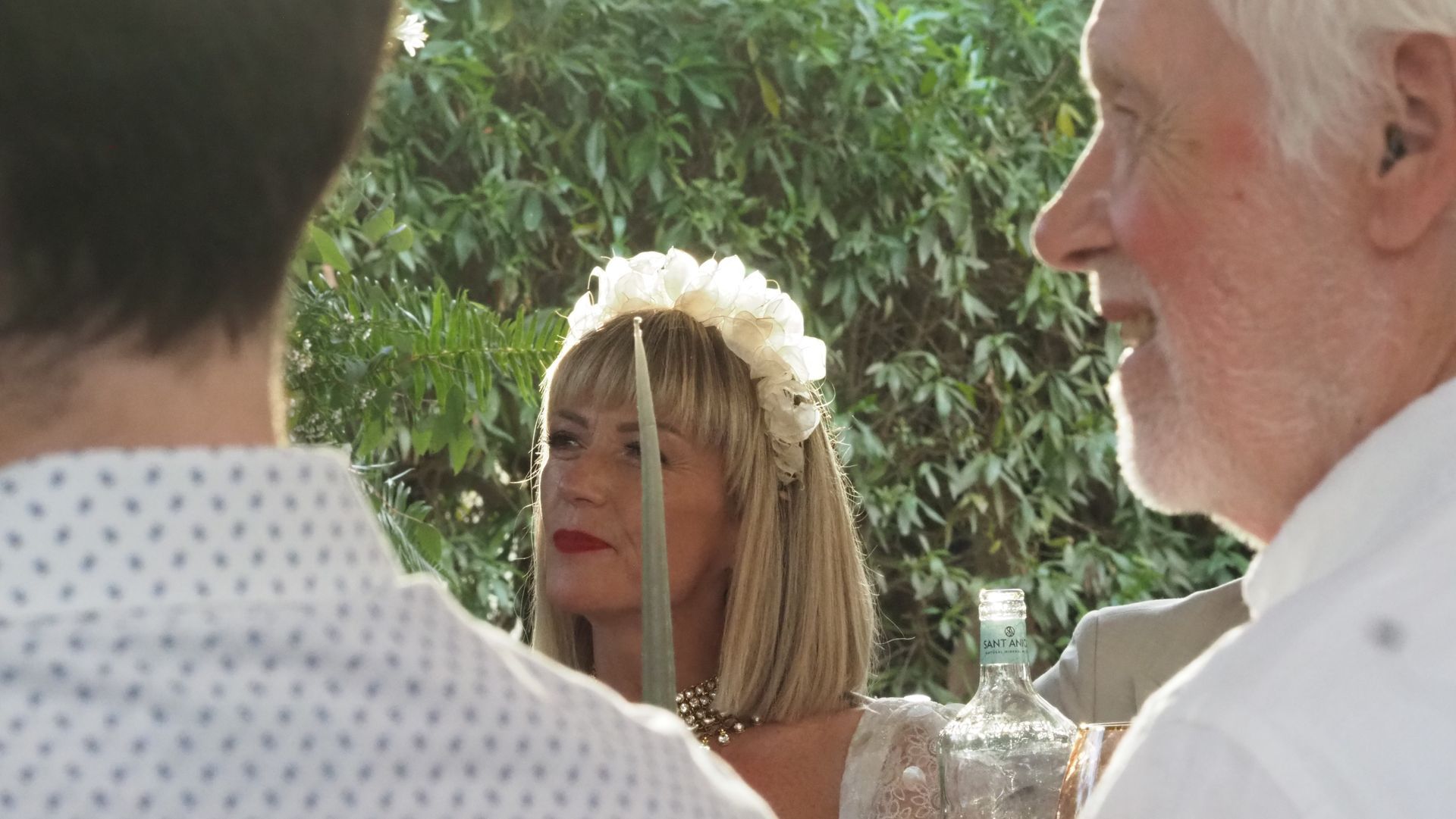 Outdoor gathering with a flower-crowned woman framed between two men in white shirts, greenery behind her