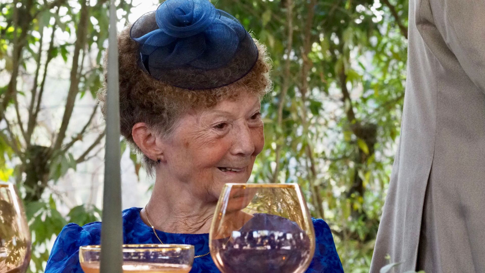 Smiling woman in a blue hat at an outdoor table with wine glasses and greenery behind her