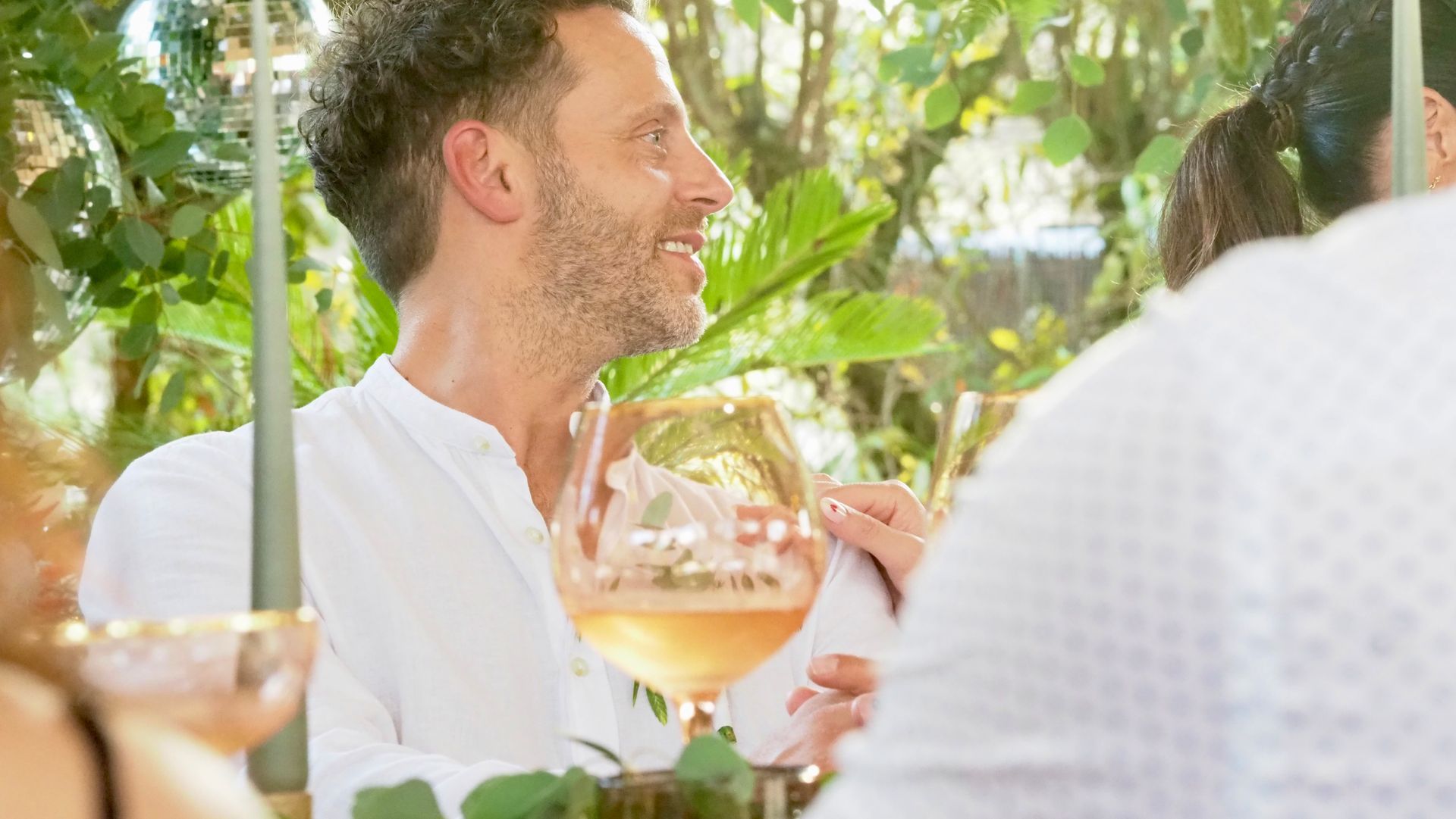 Man in white shirt smiling at an outdoor table, holding a wine glass in a bright garden setting.