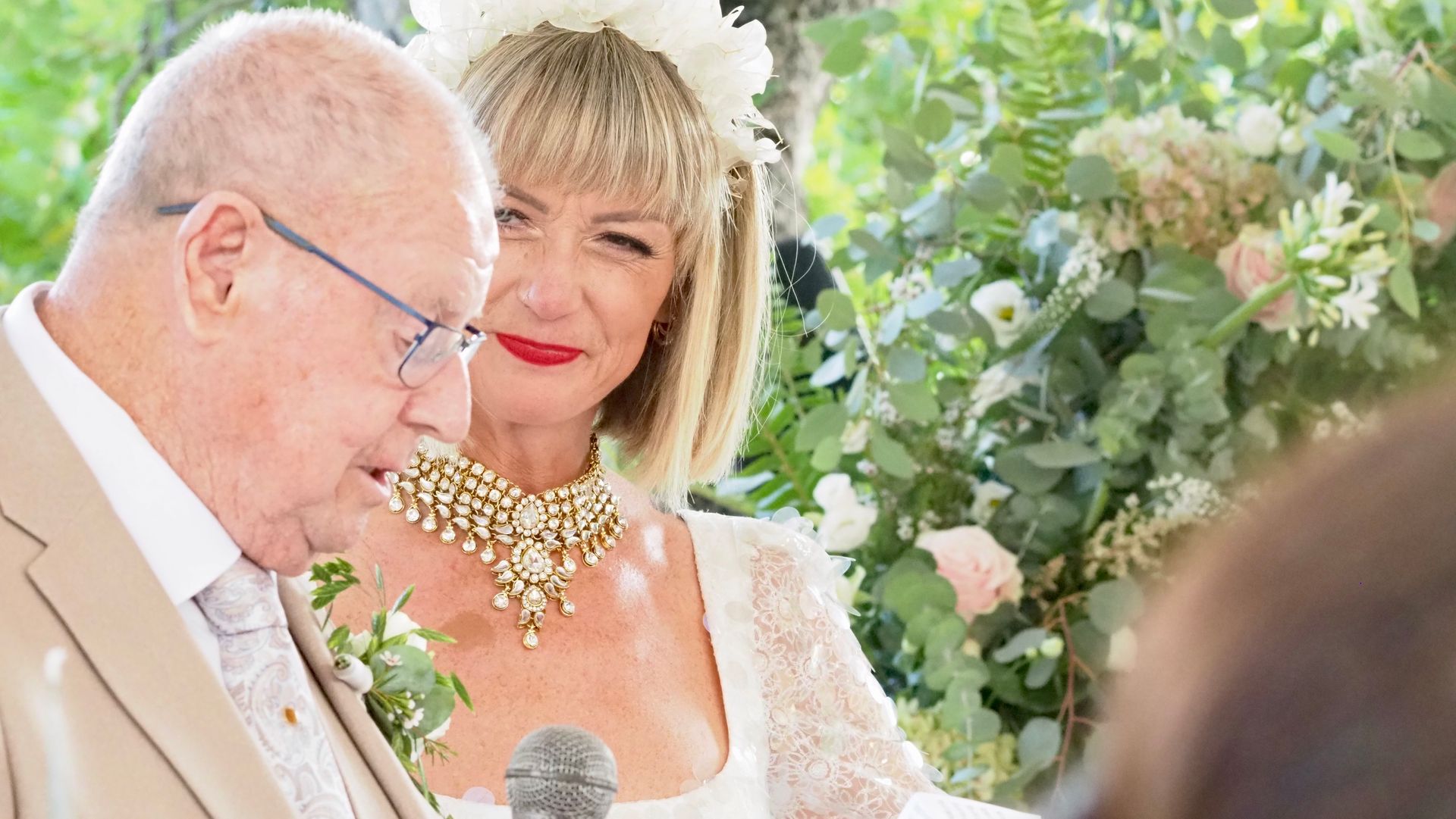 Elderly man in a light suit speaking beside a smiling woman in a white dress at a floral wedding ceremony