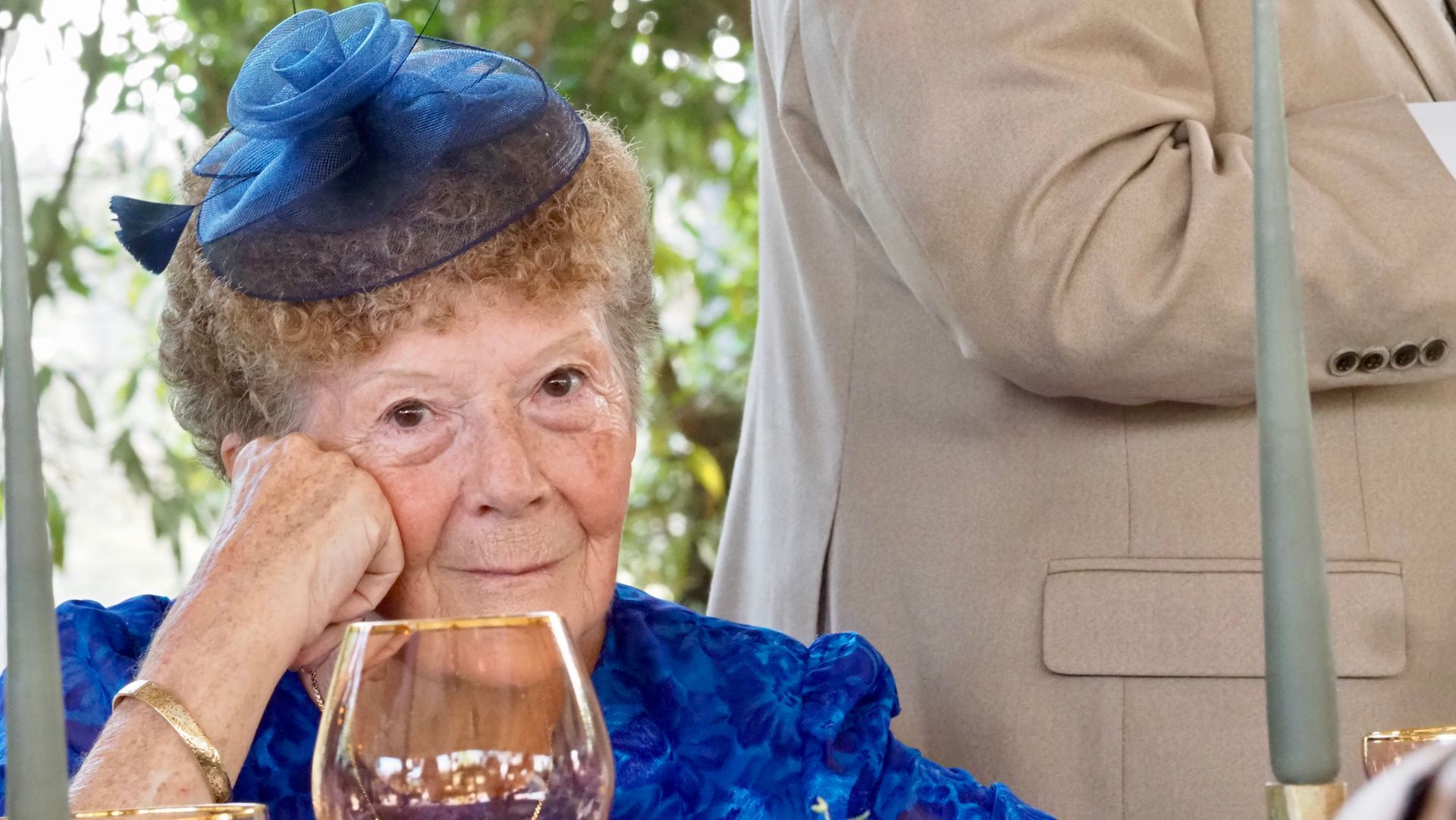 Woman in a blue hat and dress resting her chin on her hand at a formal event, with a wine glass in front.