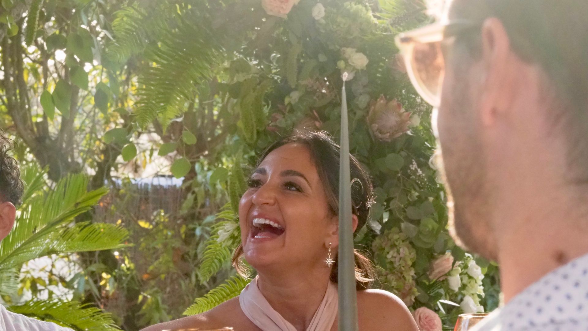 Two people smiling and talking in a sunlit garden with pink flowers and green leaves.