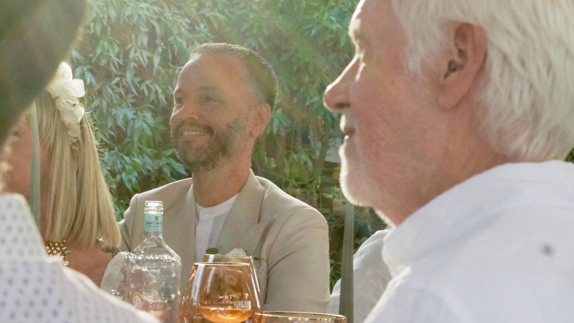 People sitting outdoors at a sunny table, smiling and talking with drinks and a water bottle nearby