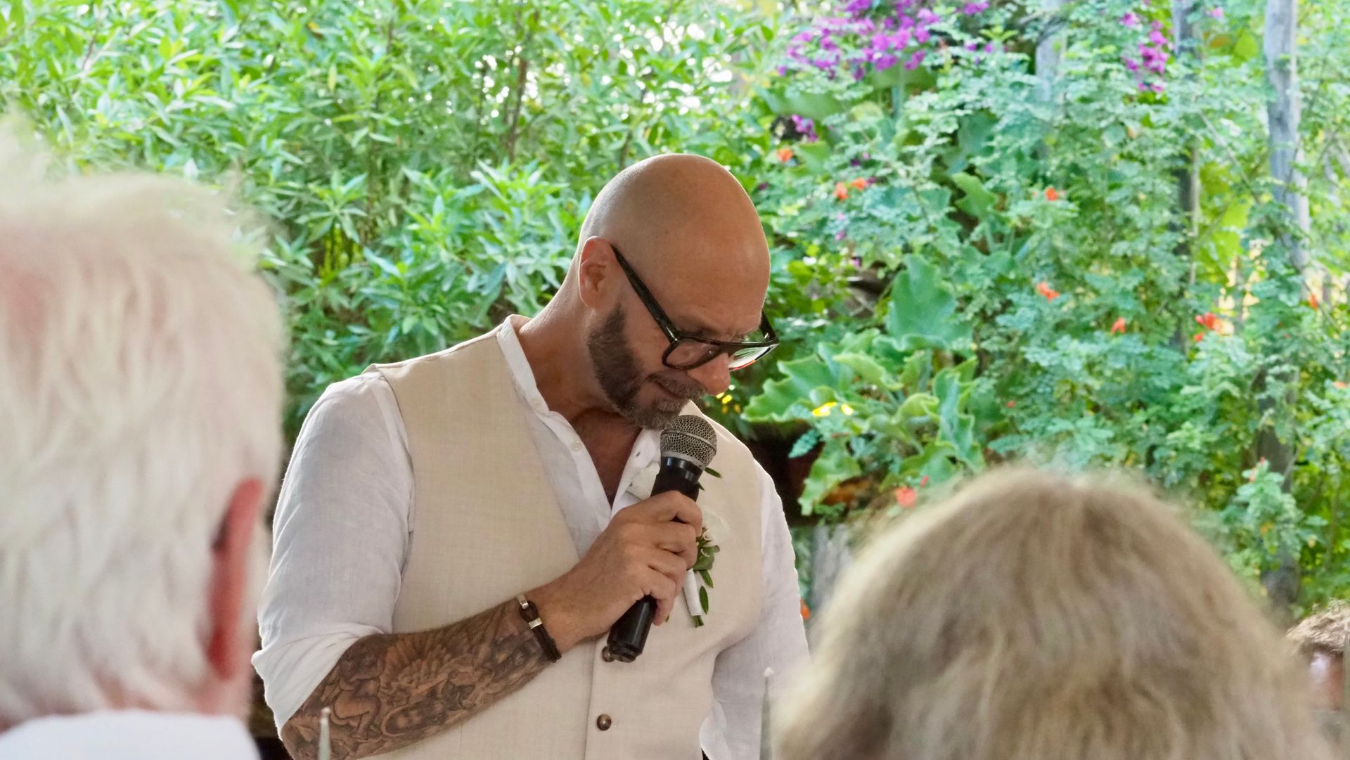 Man speaking into a microphone at an outdoor gathering, with greenery and seated audience in the background
