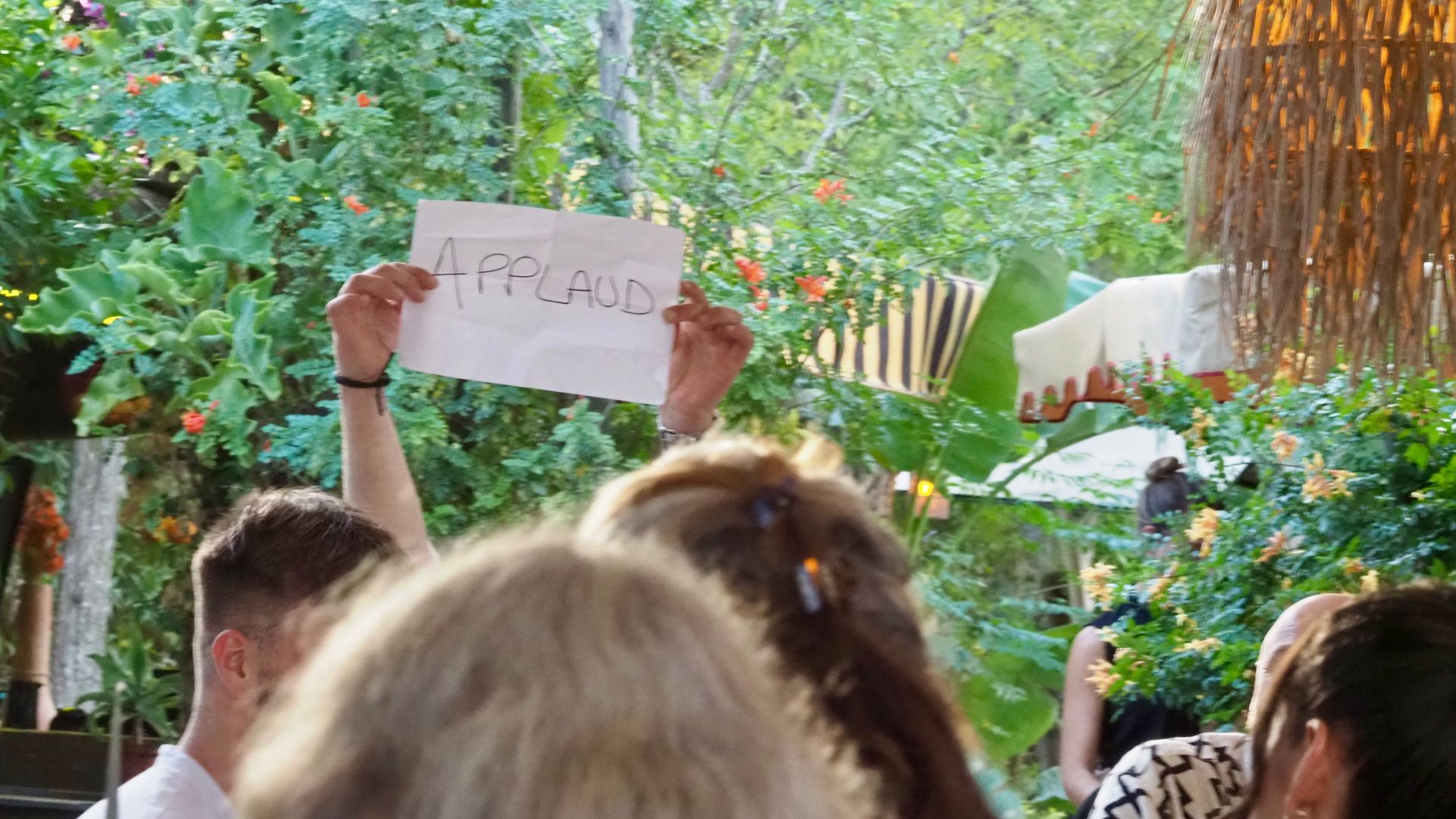 Crowd outdoors, with a person holding a hand-drawn sign above their head amid greenery.