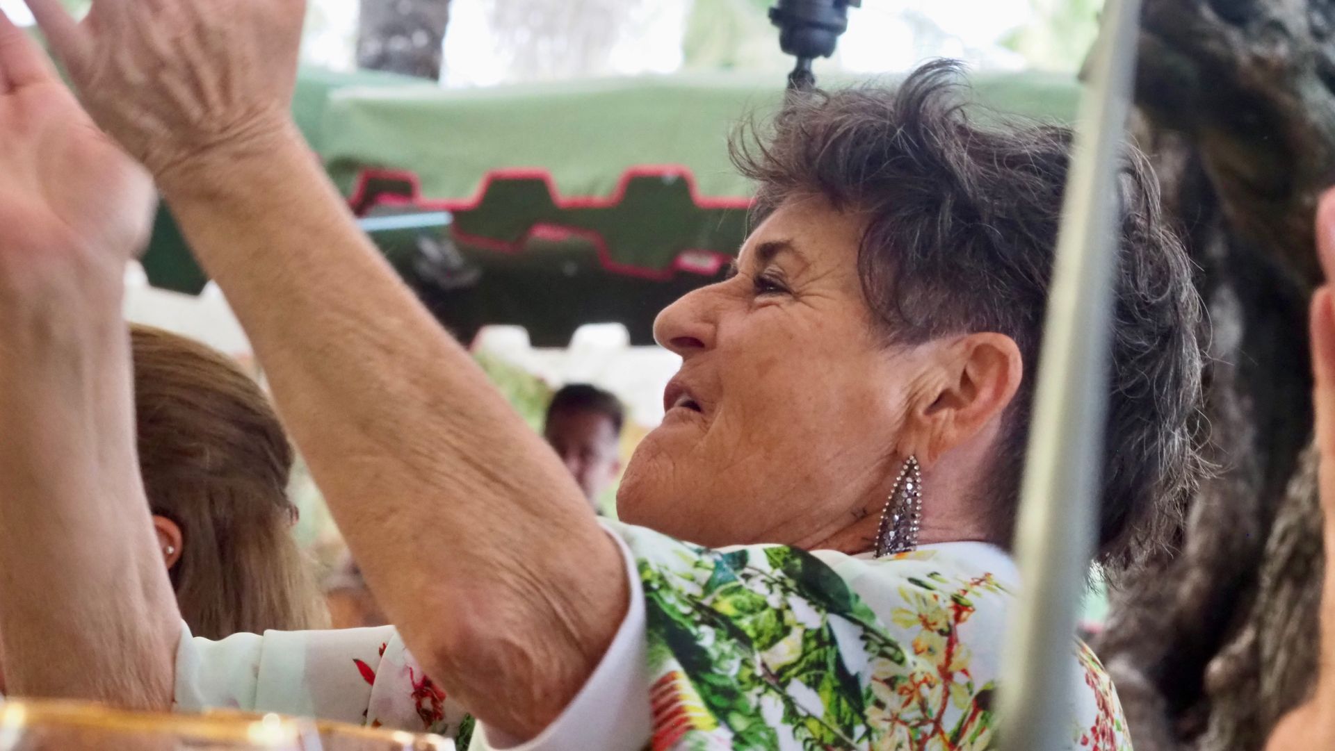 Woman in a floral shirt raising her arm at an outdoor gathering under a canopy.