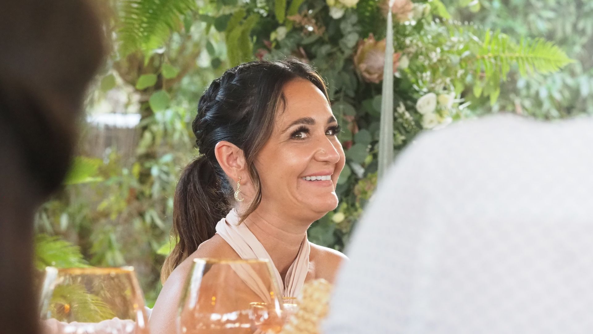 Smiling woman with dark hair sitting at a table outdoors under greenery, looking to the side
