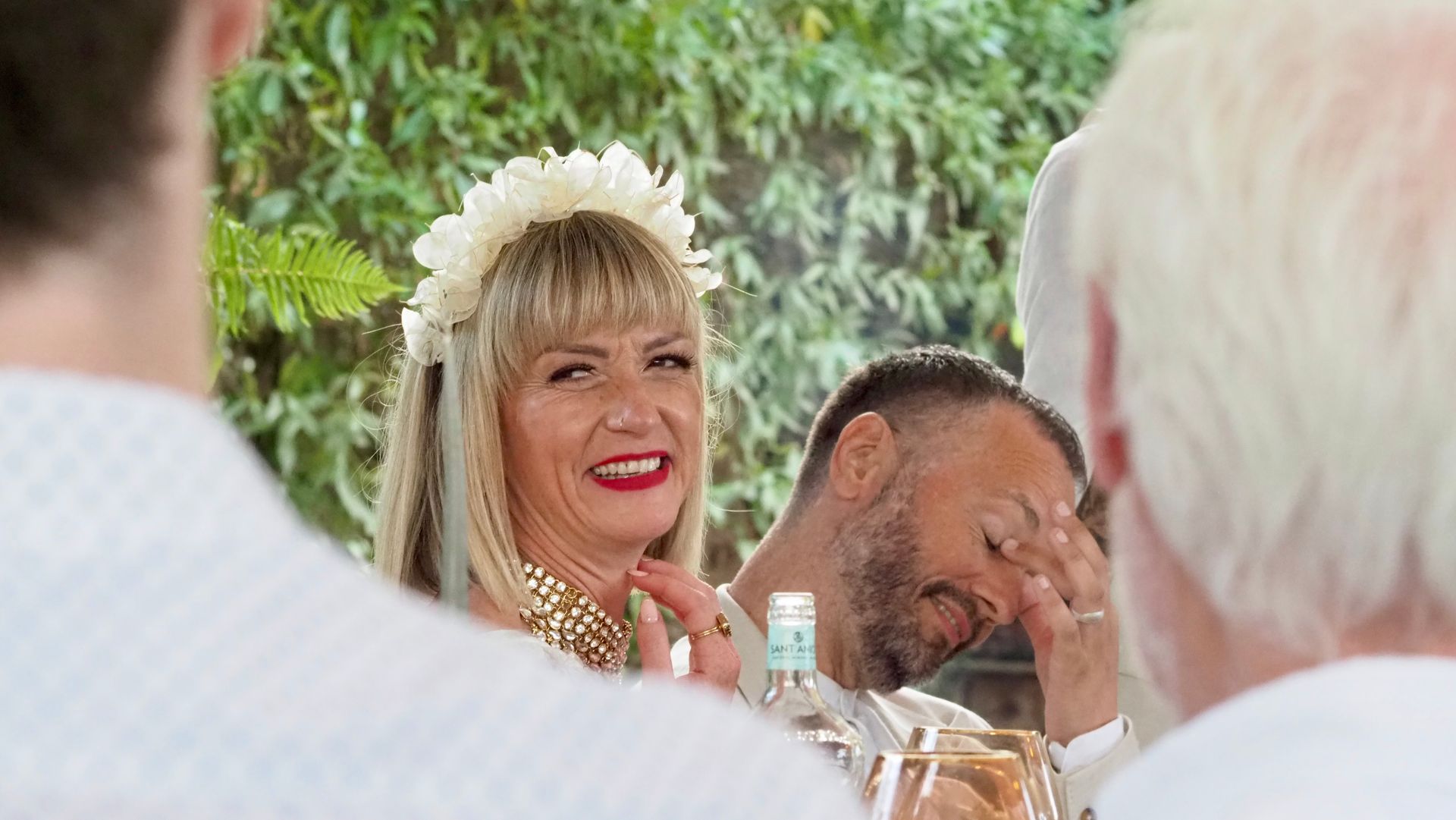 Smiling woman with red lipstick at an outdoor gathering, with a man rubbing his eyes nearby