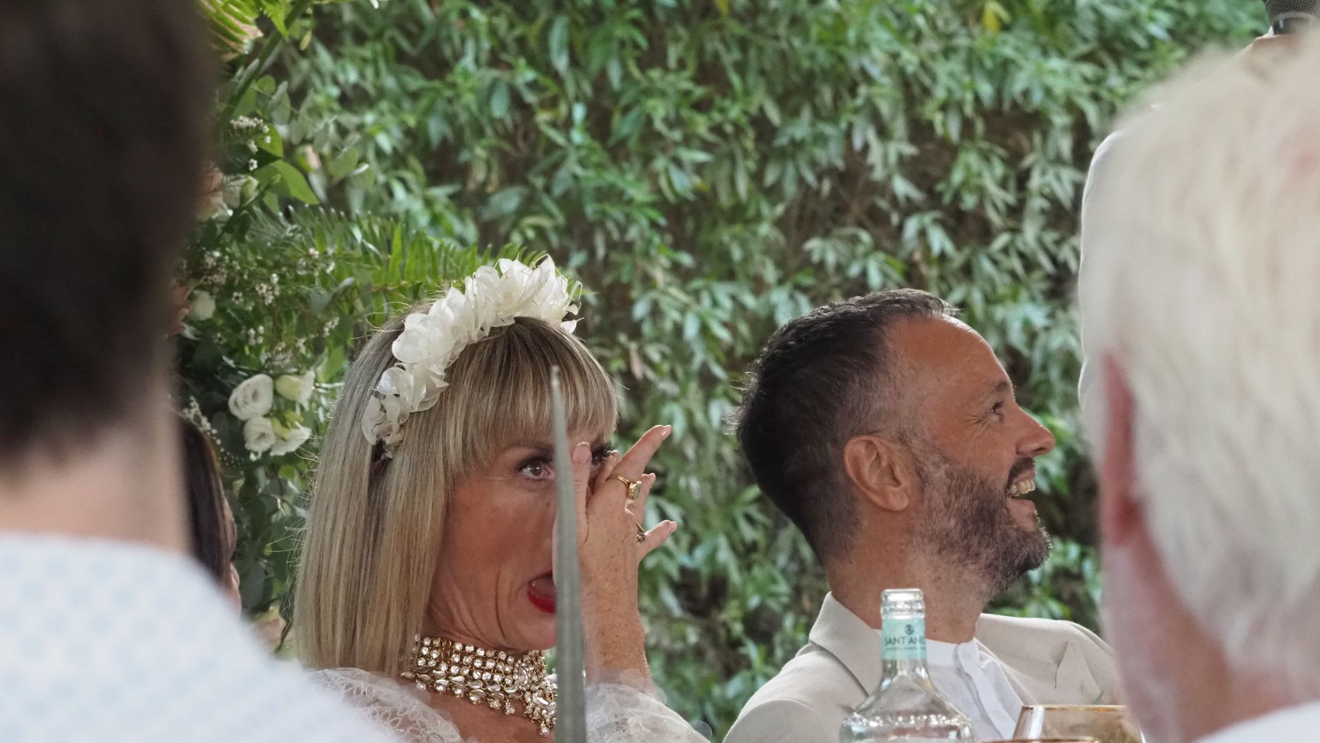 Woman in a floral headpiece and man seated at an outdoor event, with greenery in the background.